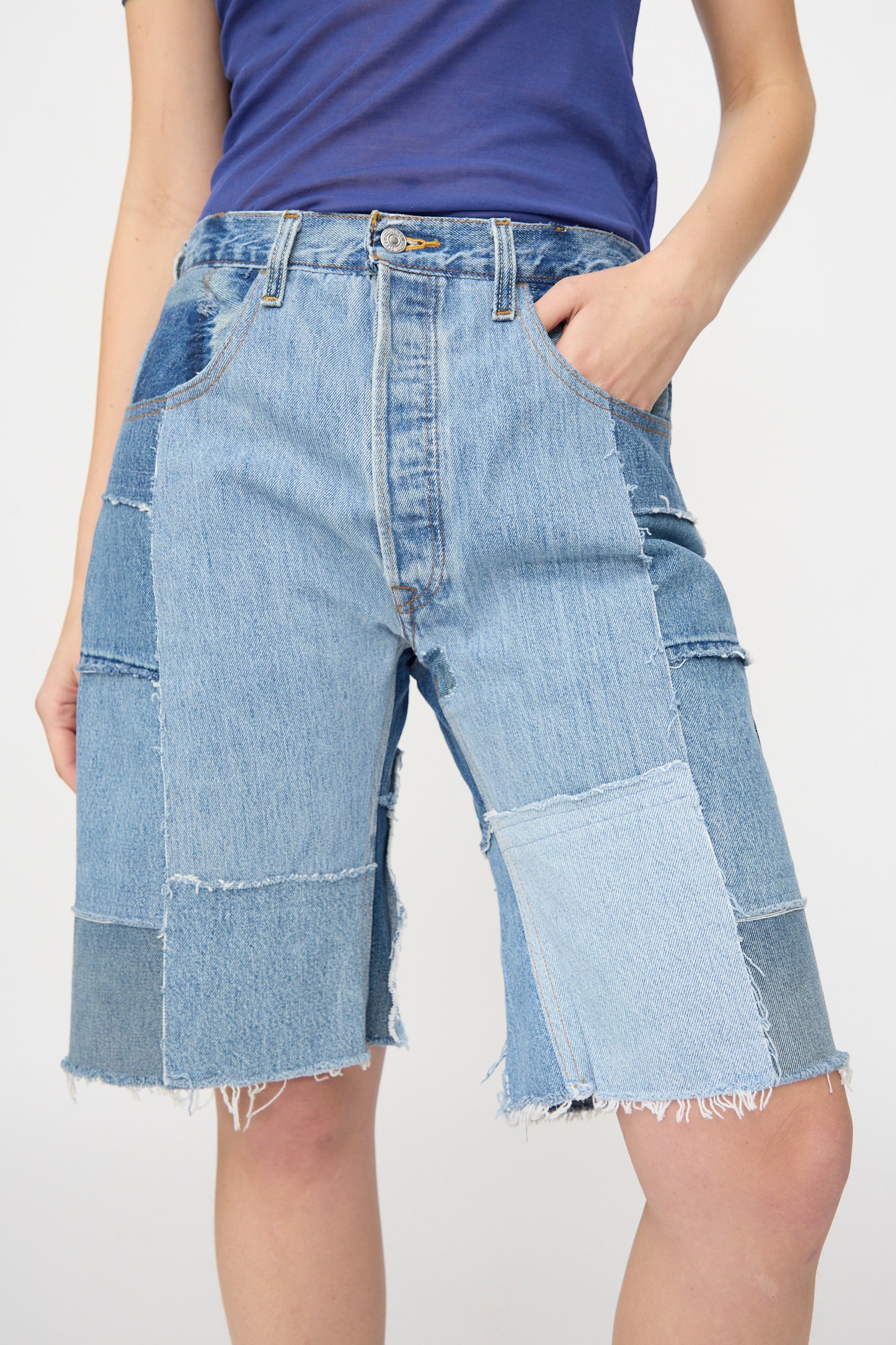 Model wears the B Sides Rework Madras Short in Vintage Indigo—relaxed fit patchwork denim shorts with frayed hems—paired with a blue top, standing with one hand in their pocket against a plain background.