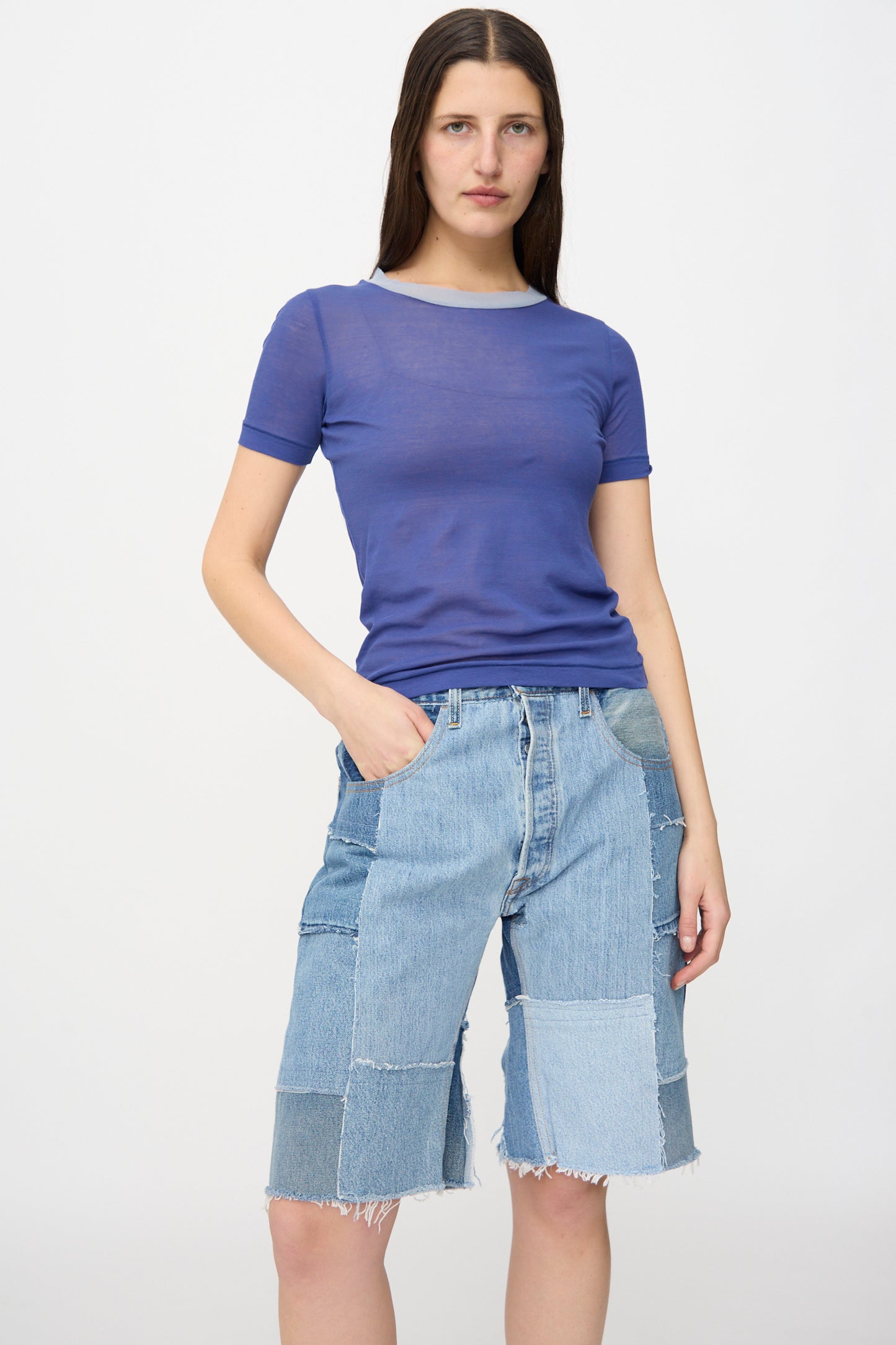 A woman stands against a plain background wearing the B Sides Rework Madras Short in Vintage Indigo, paired with a sheer blue t-shirt and one hand casually in her pocket.