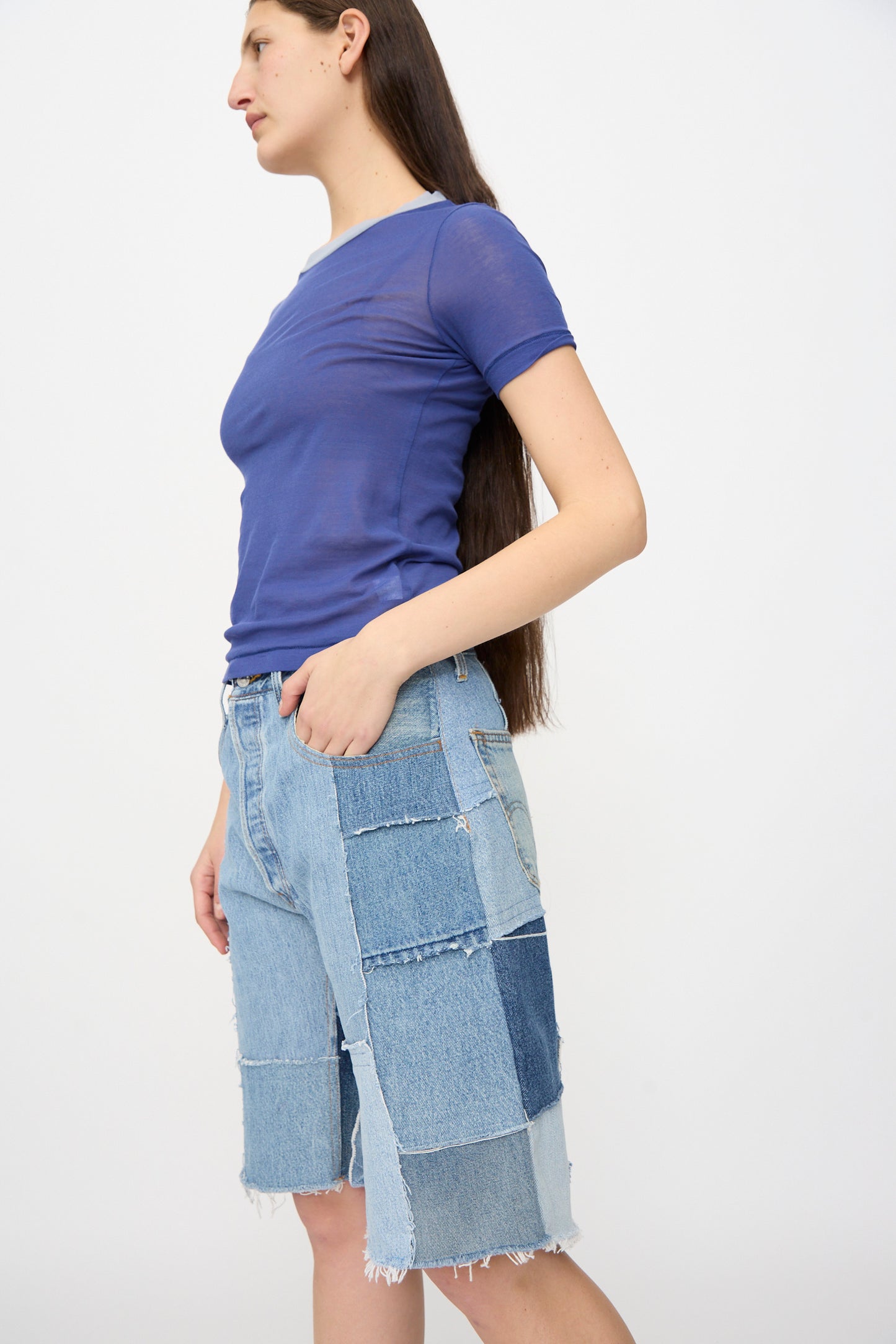 A person with long brown hair wears a sheer blue t-shirt and B Sides Rework Madras Short in Vintage Indigo, standing against a plain white background.