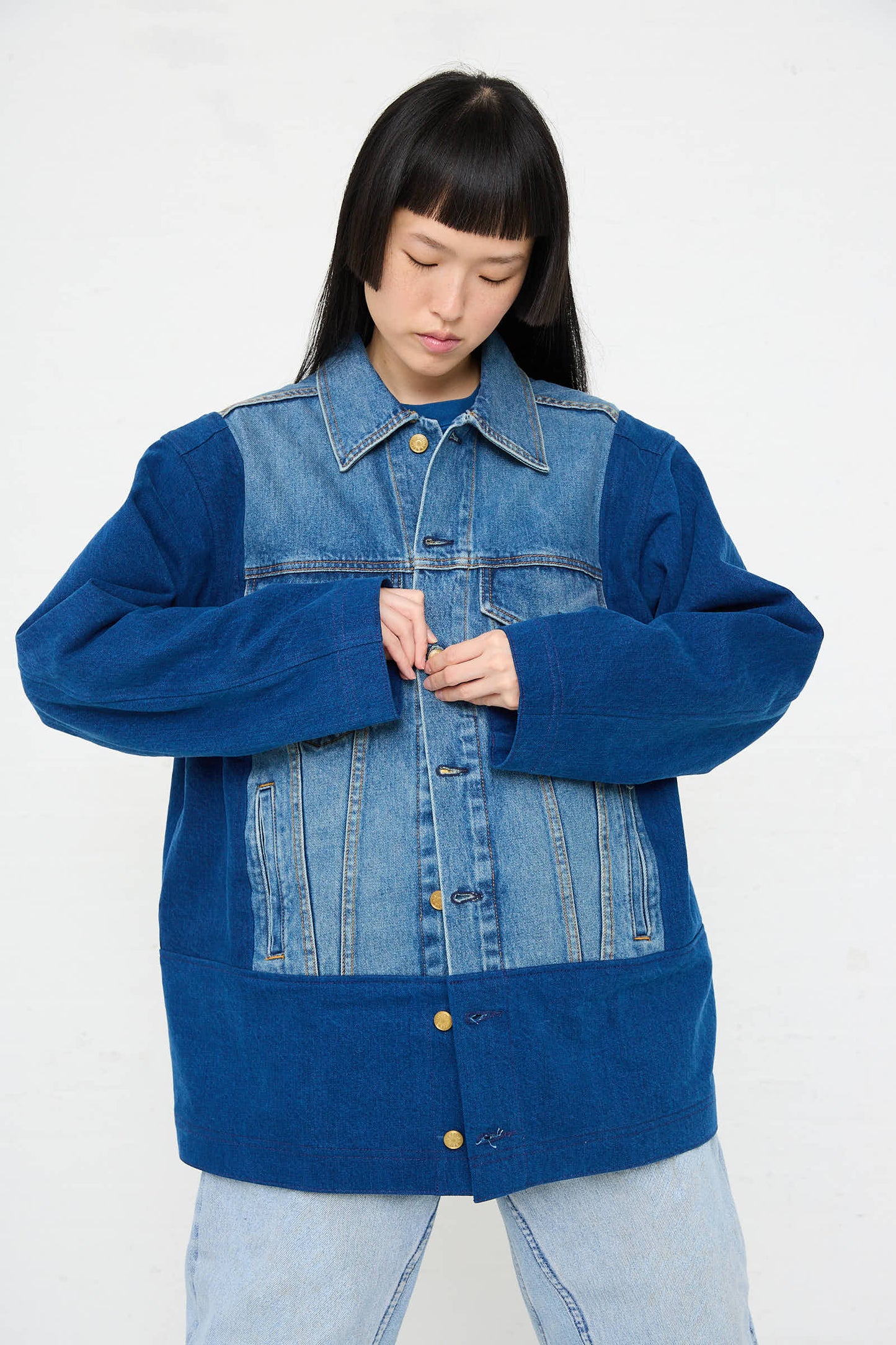 A person wearing the Reworked Barn Jacket in Quincy Blue by B Sides over a lighter, multi-colored denim shirt is seen fastening the middle button. They have long, straight hair with bangs, and they are looking down. The relaxed fit of their outfit stands out against the plain white background.