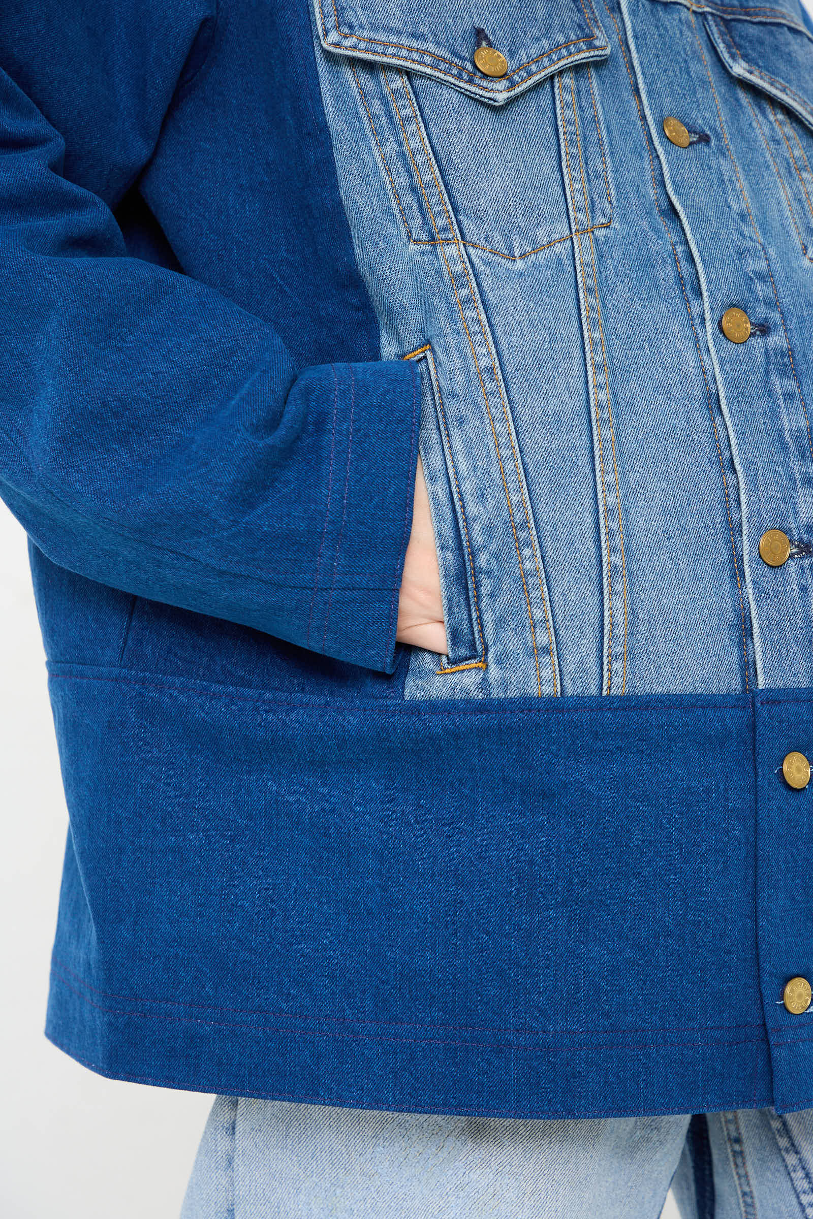 Close-up of a person wearing the B Sides Reworked Barn Jacket in Quincy Blue, featuring a relaxed fit. The left hand is tucked in a front pocket, highlighting brass button details and intricate stitching patterns. The person pairs it with light blue jeans, creating a stylish multi-color denim look.