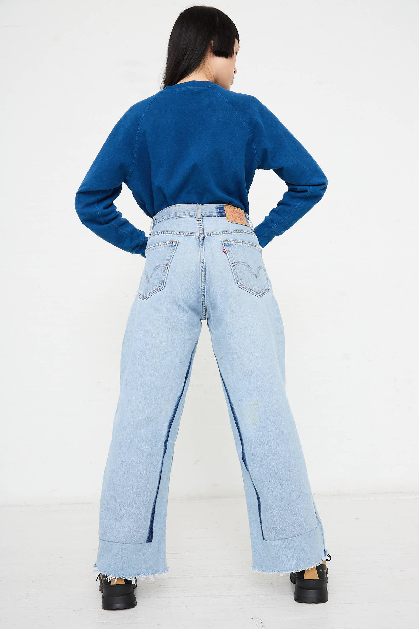 Person facing away, wearing a blue long-sleeve shirt, light blue Reworked Culottes in Vintage Indigo by B Sides with a label on the waist, and black shoes. The background is plain white.