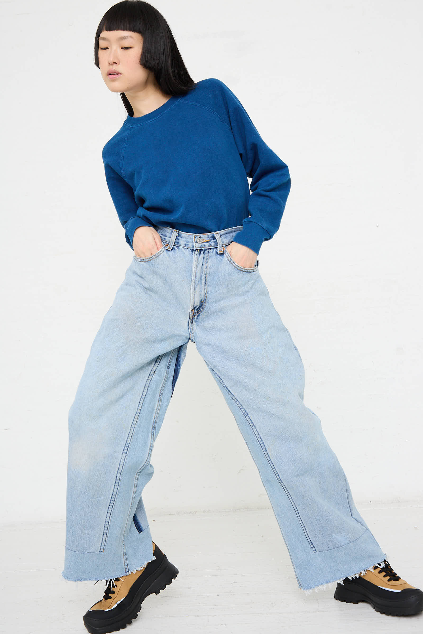 A person wearing a blue sweatshirt and B Sides Reworked Culotte in Vintage Indigo, with their hands in their pockets, stands against a white background. They are also sporting tan and black boots.