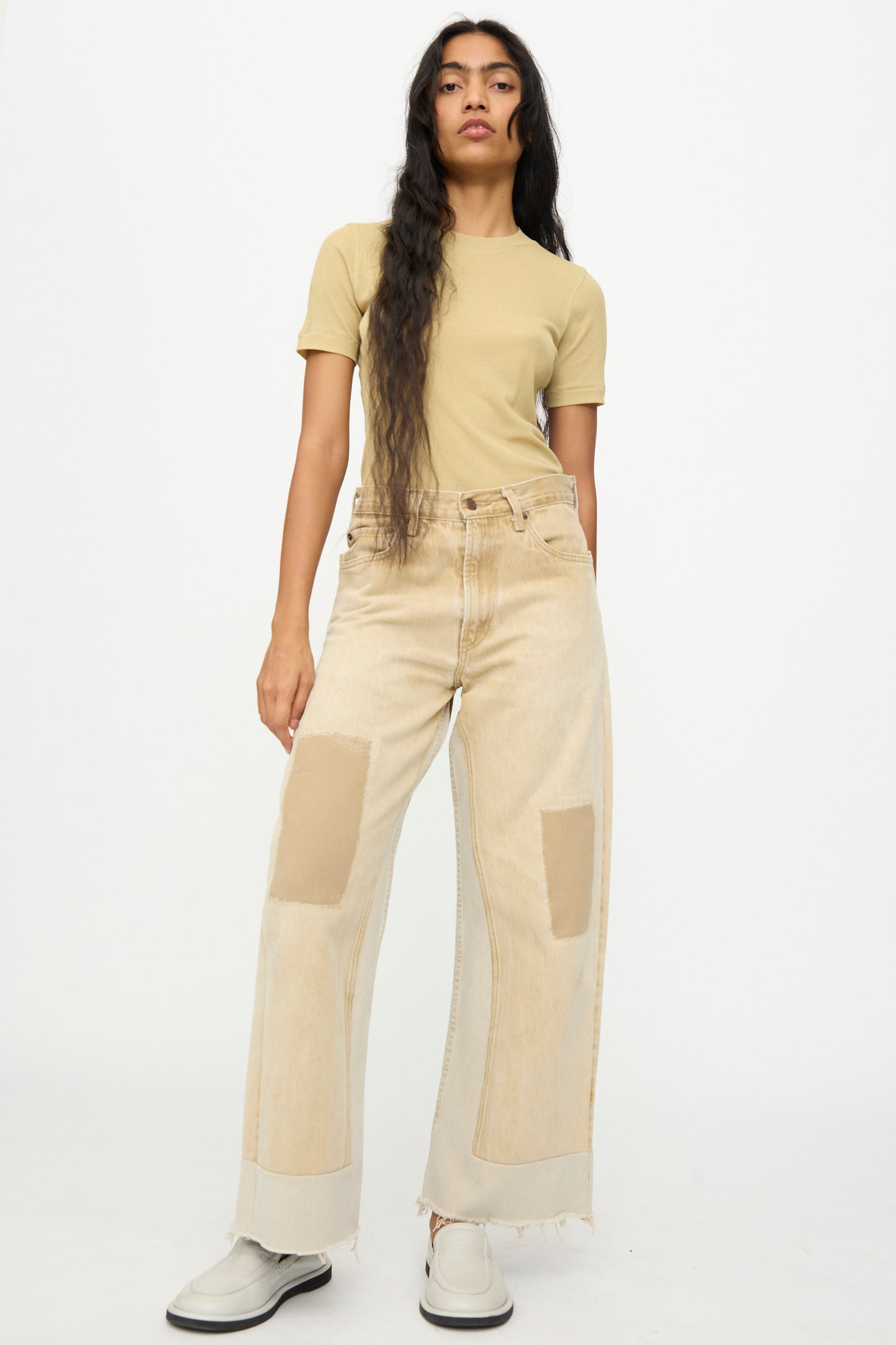 A person stands against a plain white background wearing a beige short-sleeve shirt, B Sides Reworked Vintage Culotte in Khaki, and white shoes.
