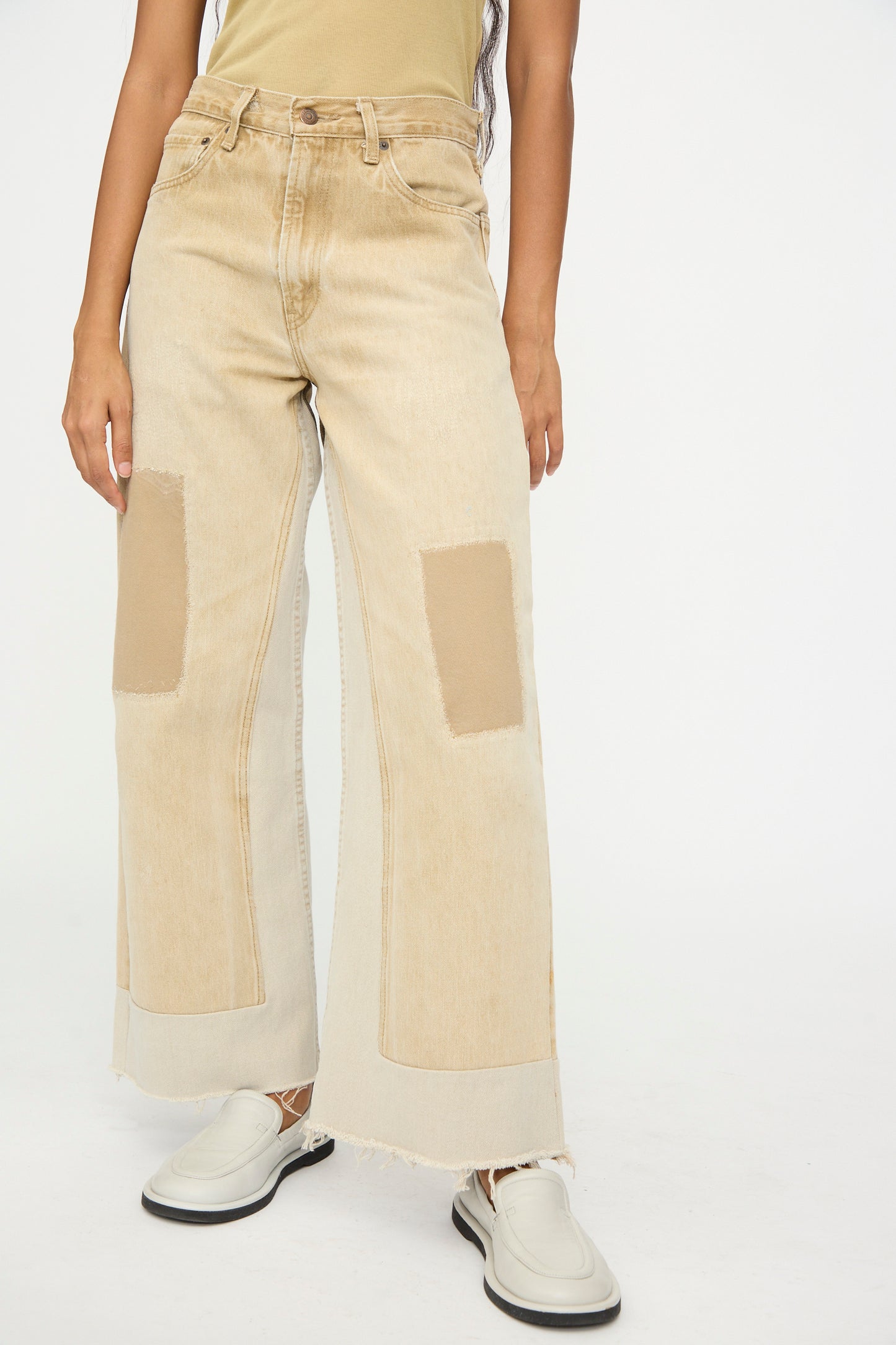 A person wears the B Sides Reworked Vintage Culotte in Khaki—relaxed fit, wide-leg jeans with knee patches and frayed hems—paired with a tan top and white slip-ons, standing against a plain background.
