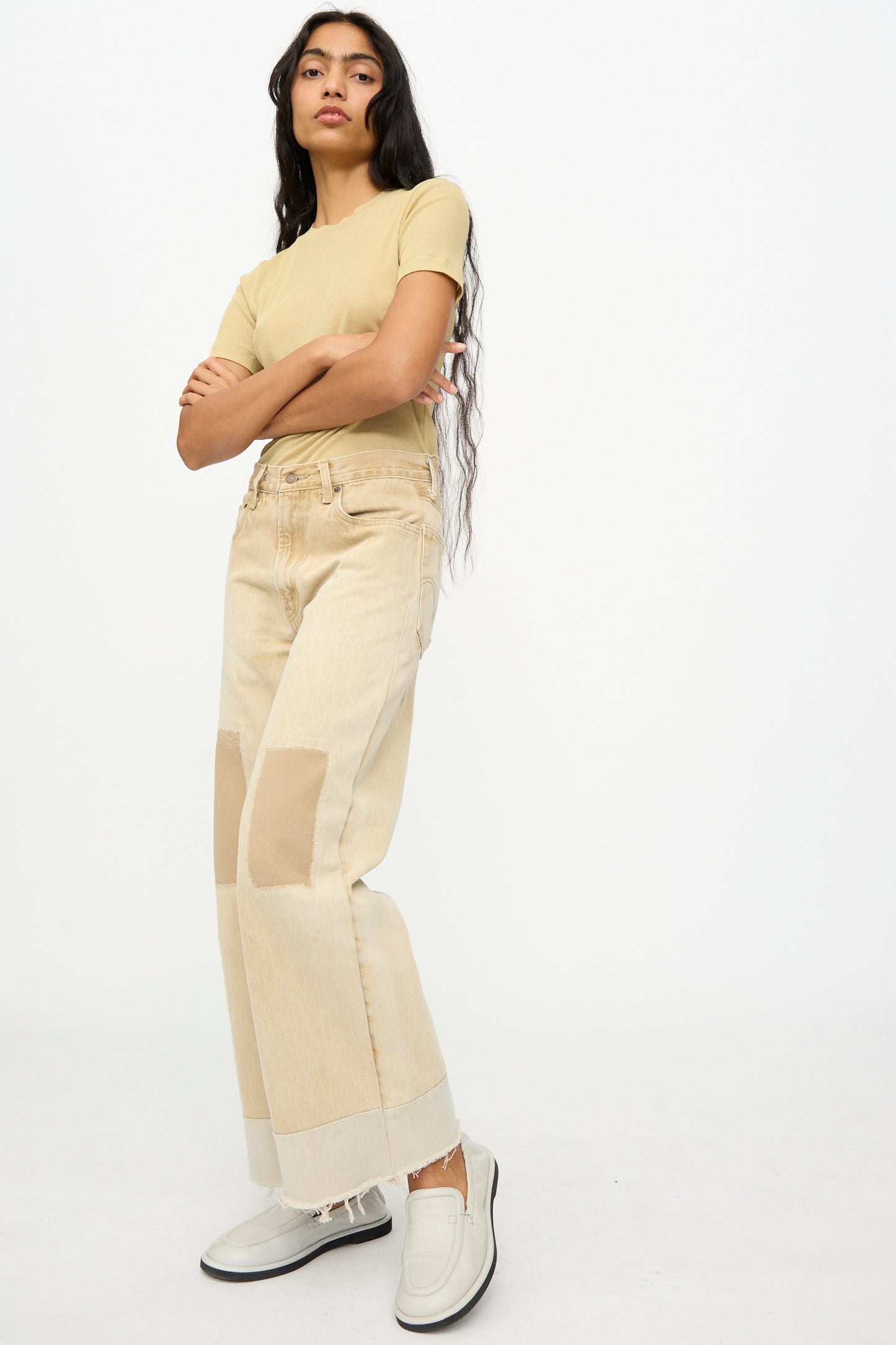 Person standing with arms crossed, wearing a beige t-shirt, B Sides Reworked Vintage Culotte in Khaki, and white slip-on shoes against a plain white background.