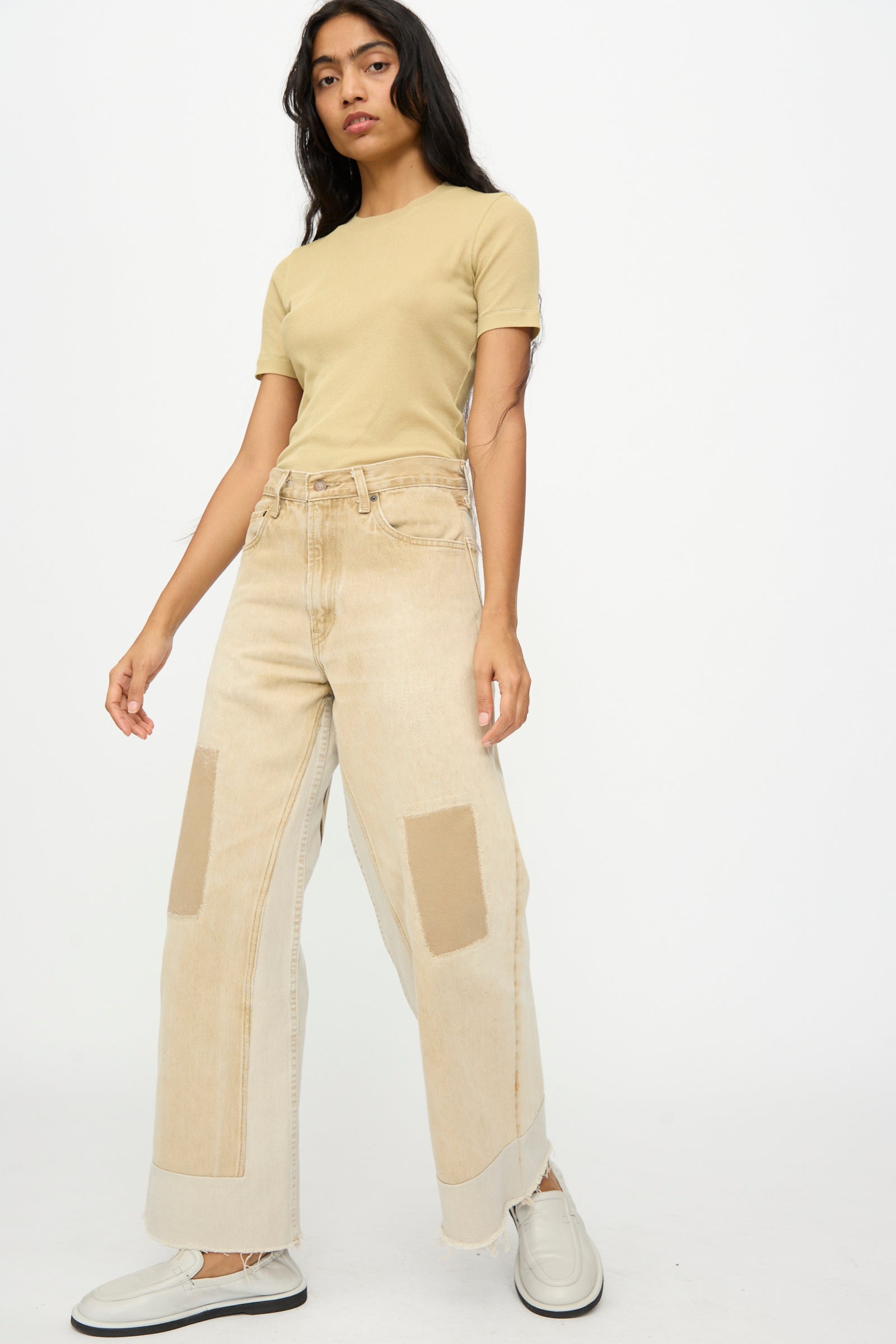A woman stands against a plain background wearing a beige t-shirt, B Sides Reworked Vintage Culotte in Khaki, and light-colored slip-on shoes.