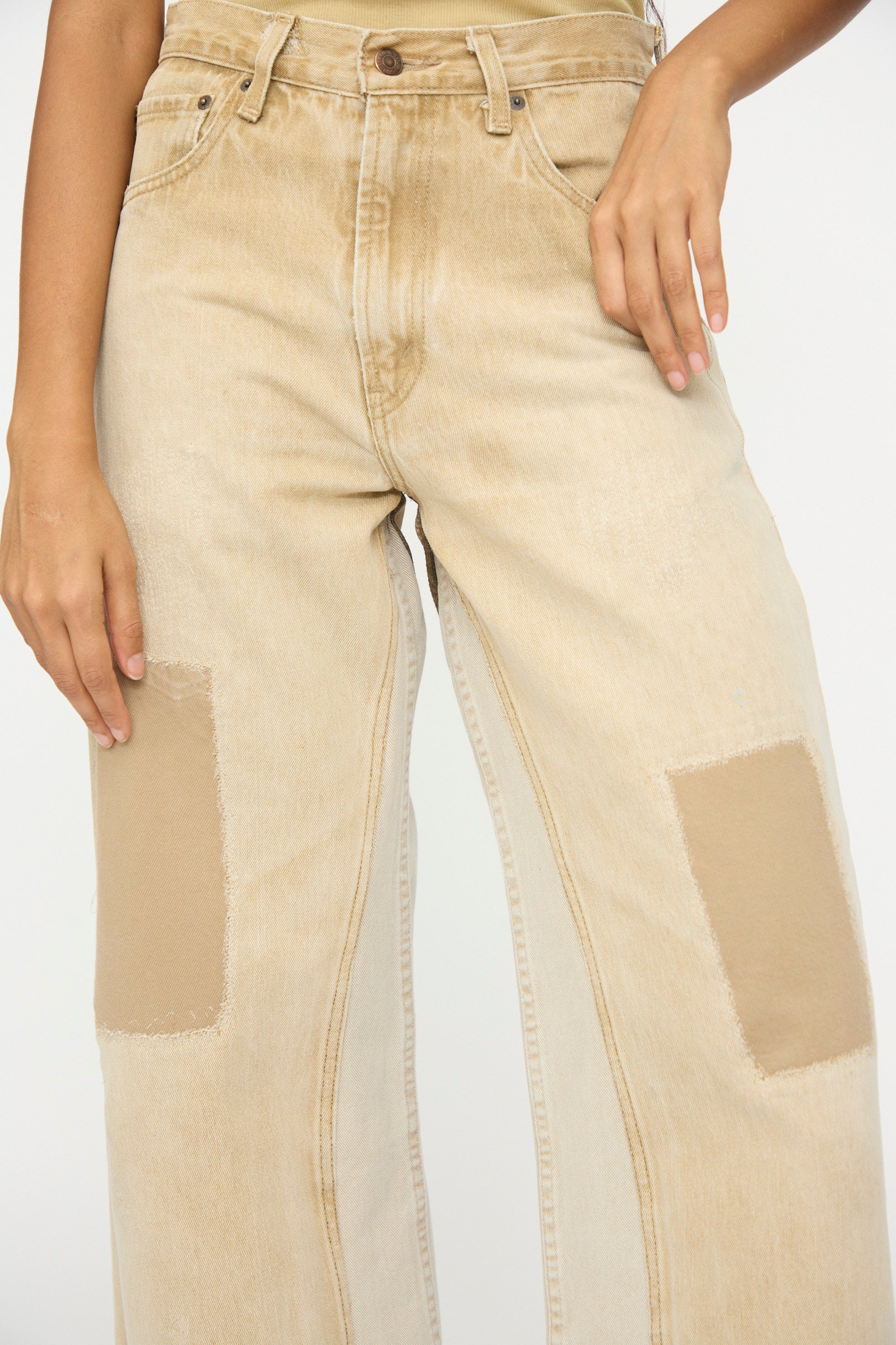 A person wears B Sides’ Reworked Vintage Culotte in Khaki—relaxed-fit, mid-rise denim jeans with large tan thigh patches—standing against a plain background.