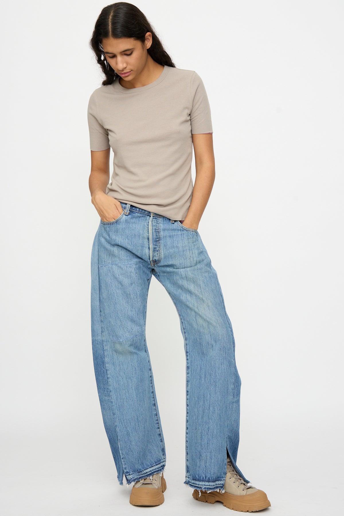 Person wearing a beige short-sleeve t-shirt, Reworked Vintage Open Hem Slouch in Indigo by B Sides, and tan shoes stands against a white background, looking down with one hand in pocket.