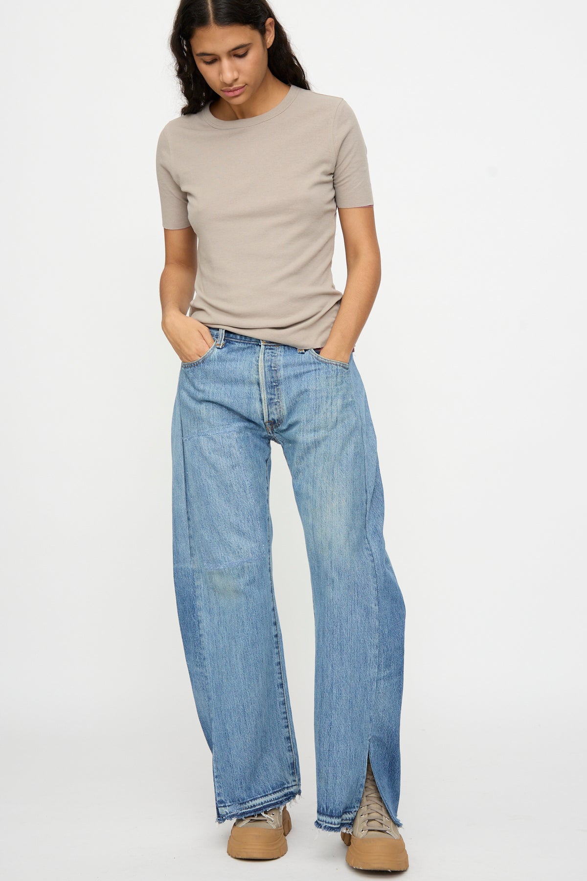 A person stands against a white background wearing a short-sleeve beige shirt, tan shoes, and B Sides Reworked Vintage Open Hem Slouch in Indigo jeans, looking down with hands in pockets.