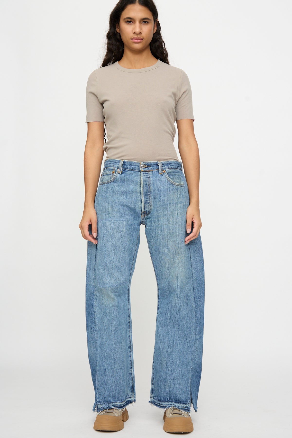 A person stands facing forward, wearing a beige short-sleeve shirt, B Sides Reworked Vintage Open Hem Slouch in Indigo jeans, and tan shoes against a plain white background.