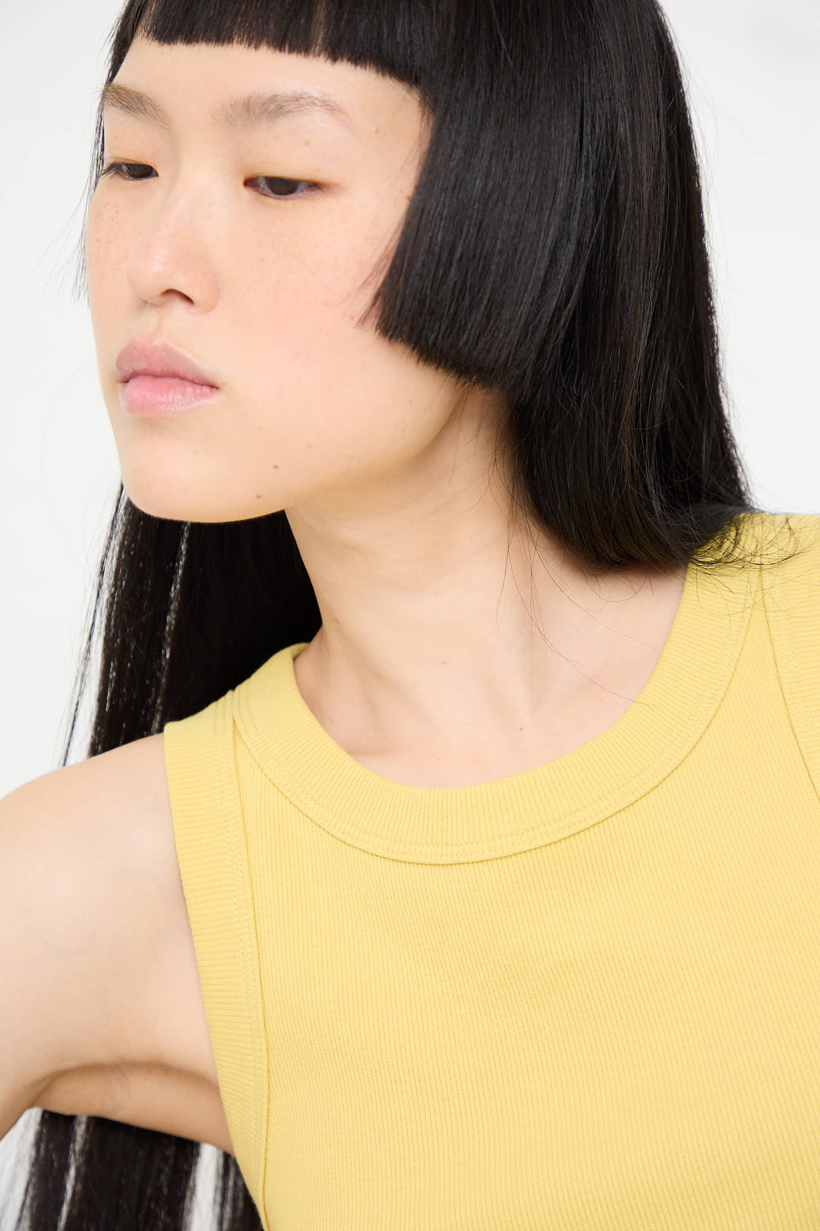 A woman with long black hair and blunt bangs wears the sleeveless Rib Tank in Daphne Overdye by B Sides, looking slightly to the side, showcasing its slim fit appeal.