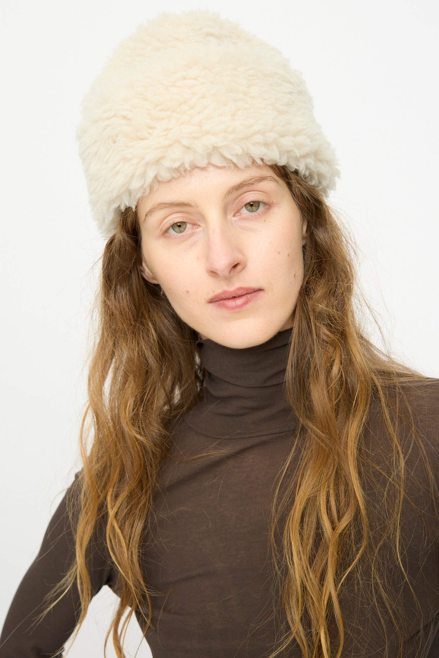 A person with long, wavy hair models the B Sides Sherpa Hat in Ivory and a sheer brown turtleneck against a plain white background.
