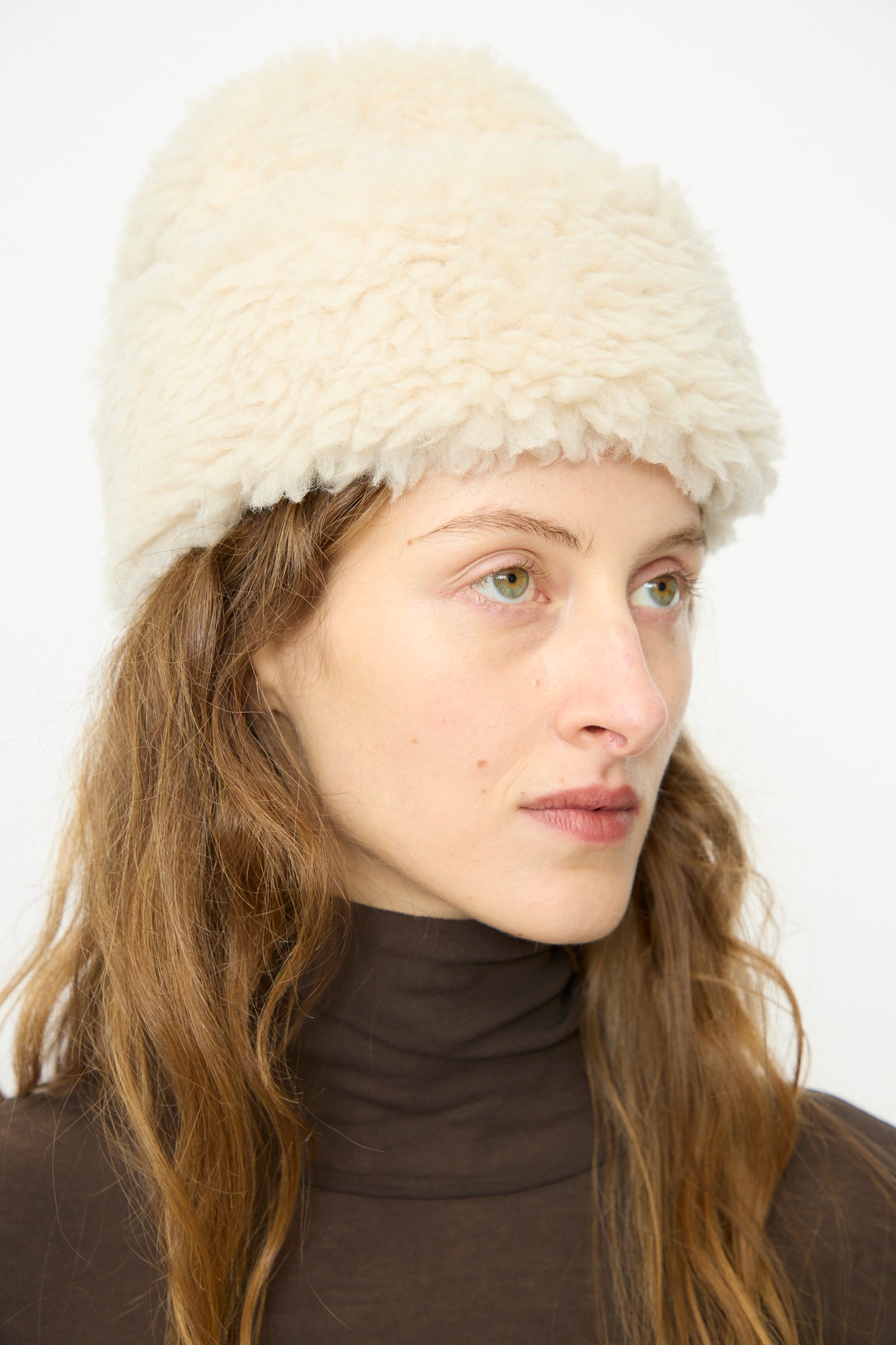 A person with long brown hair wearing a B Sides Sherpa Hat in Ivory and a dark brown turtleneck, looking slightly to the side against a plain background.