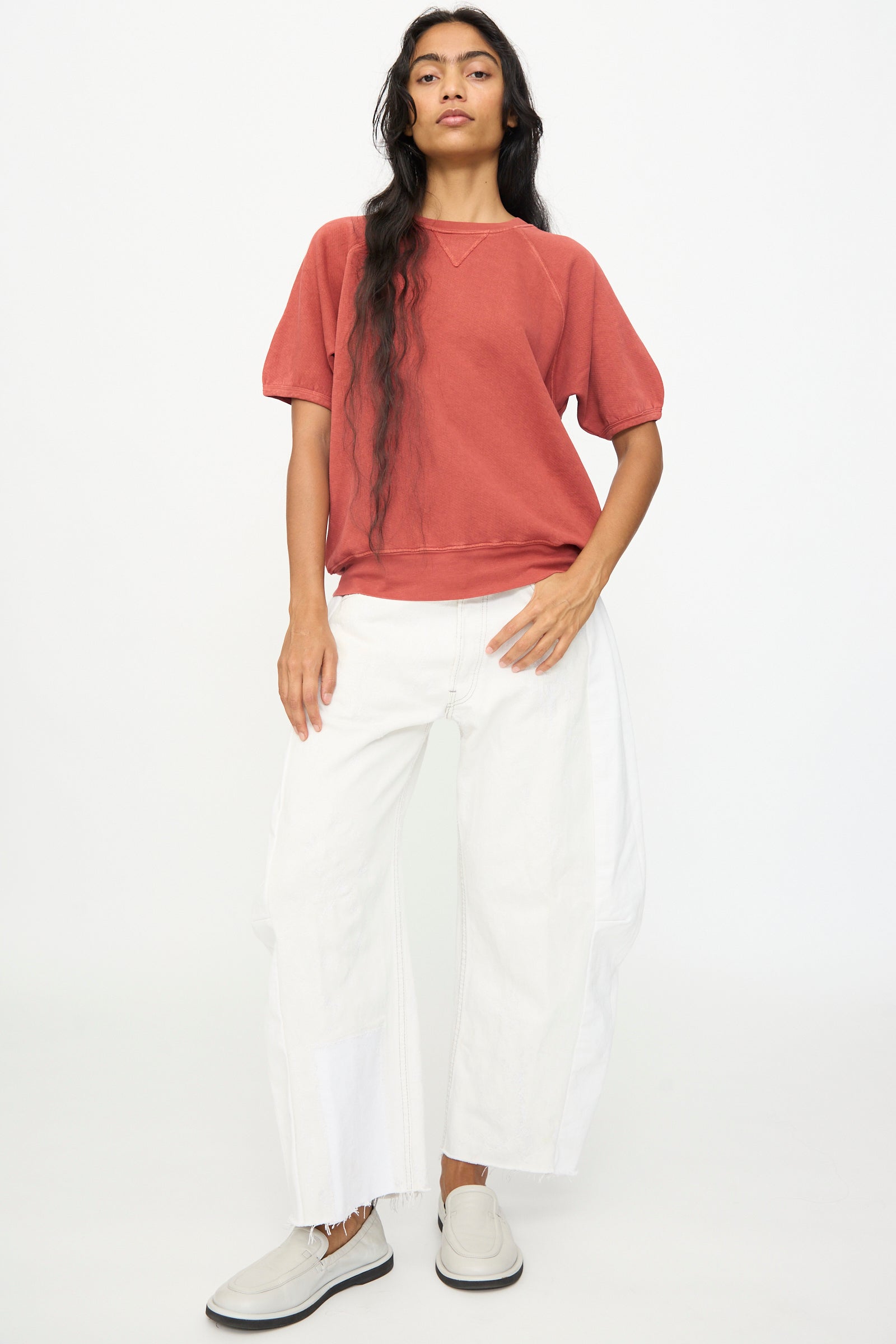 A person stands against a plain white background, wearing the B Sides Short Sleeve Sweatshirt in Faded Tomato Overdye, paired with white wide-leg pants featuring raw hems and white slip-on shoes.