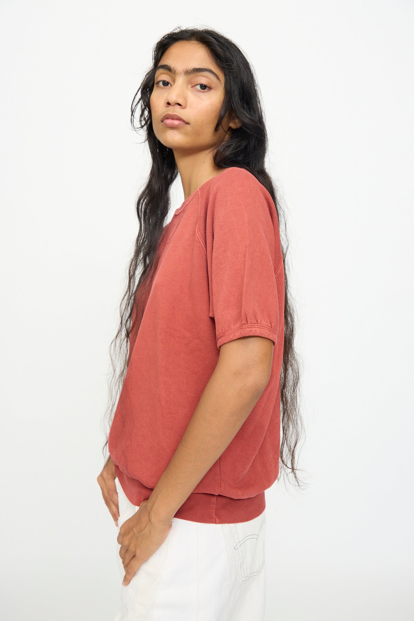 A person with long dark hair is wearing the B Sides Short Sleeve Sweatshirt in Faded Tomato Overdye and white pants, standing sideways against a plain white background.