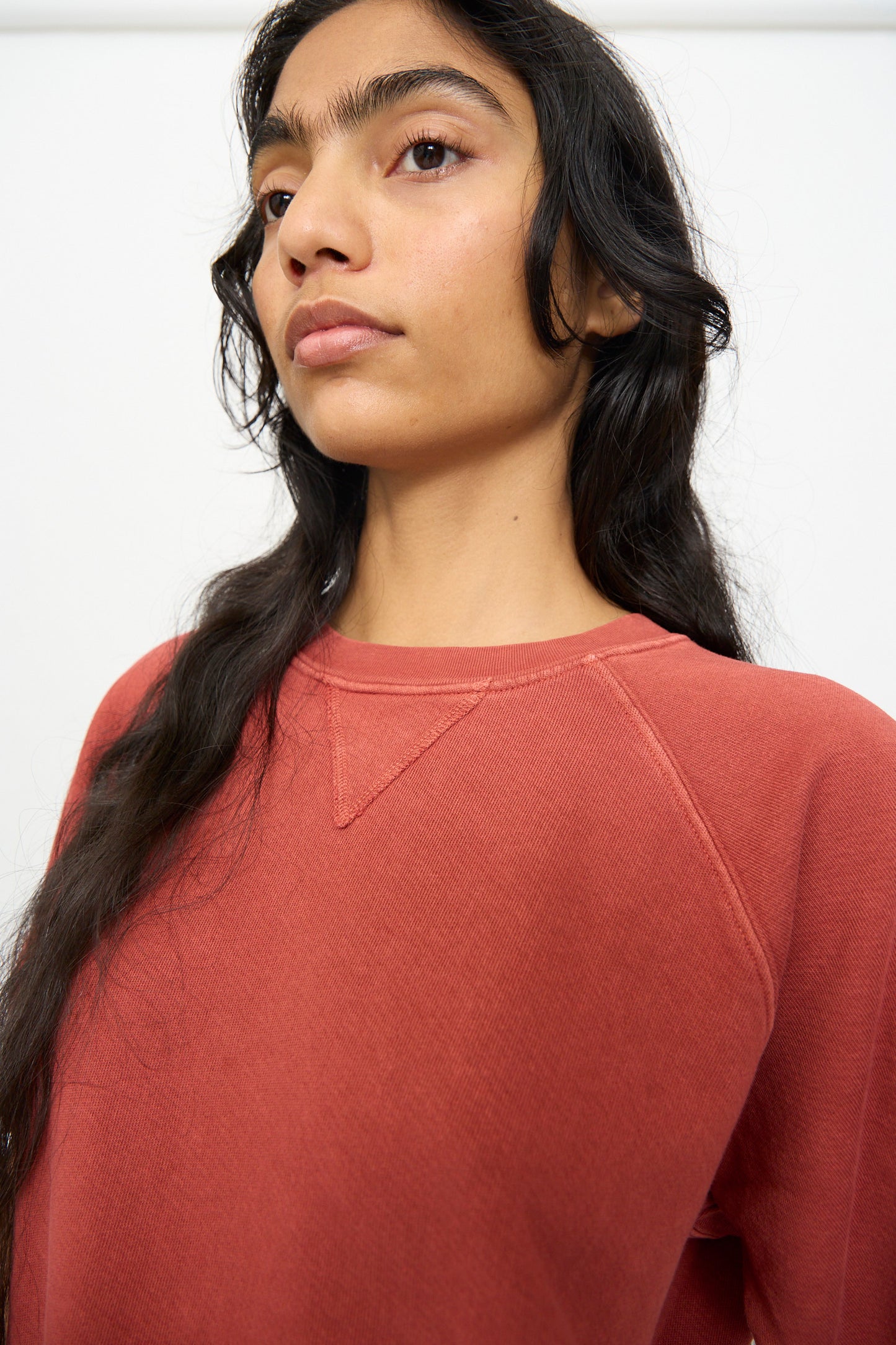 A woman with long dark hair wears the B Sides Short Sleeve Sweatshirt in Faded Tomato Overdye, styled in a relaxed fit as she gazes slightly away from the camera against a plain background.