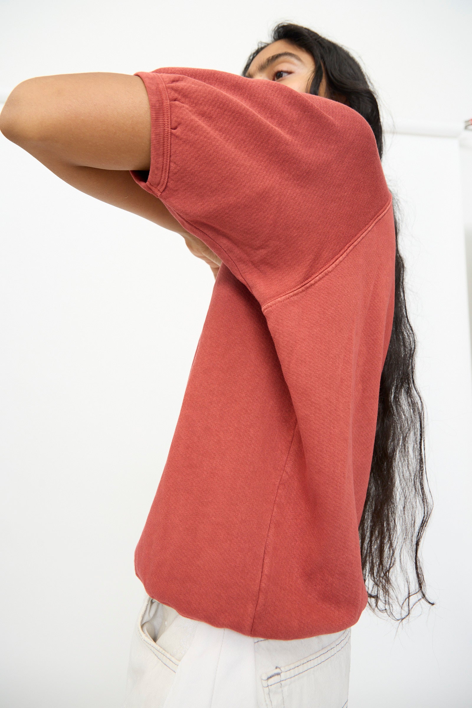 A person with long dark hair wears the B Sides Short Sleeve Sweatshirt in Faded Tomato Overdye, styled in a relaxed fit with light pants. Their face is partially obscured by their arm as they stand against a plain white background.