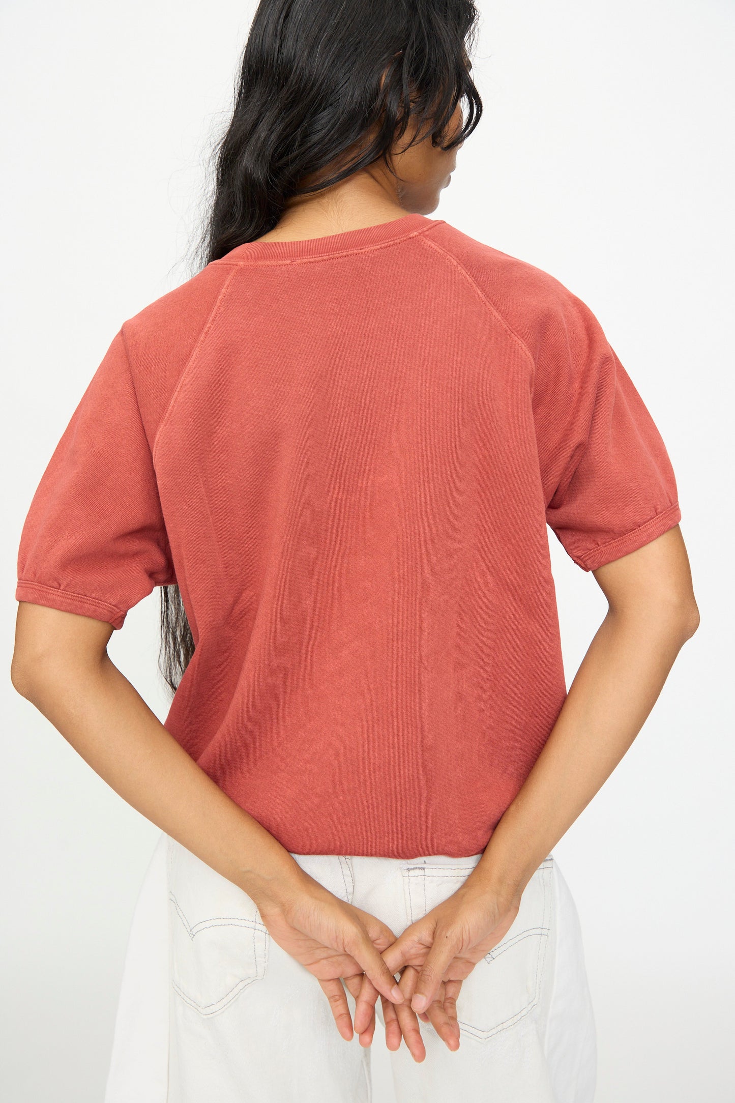A person with long dark hair stands facing away, wearing the B Sides Short Sleeve Sweatshirt in Faded Tomato Overdye—a relaxed fit French terry top—paired with white pants, hands clasped behind their back against a plain light background.