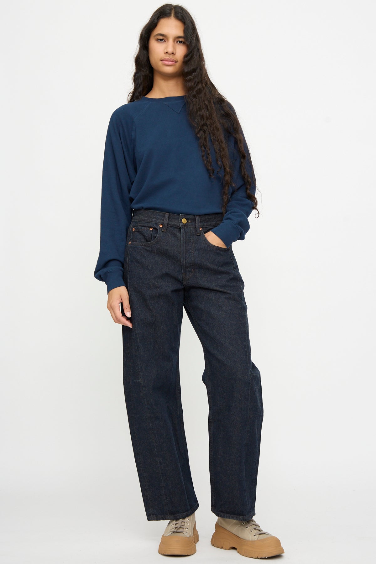 Person standing against a plain background, wearing a navy long-sleeve shirt, B Sides Slim Lasso in Indigo Brown Rinse, and beige sneakers.