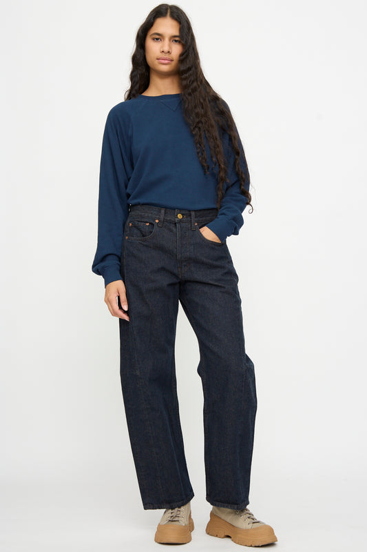 Person standing against a plain background, wearing a navy long-sleeve shirt, B Sides Slim Lasso in Indigo Brown Rinse, and beige sneakers.
