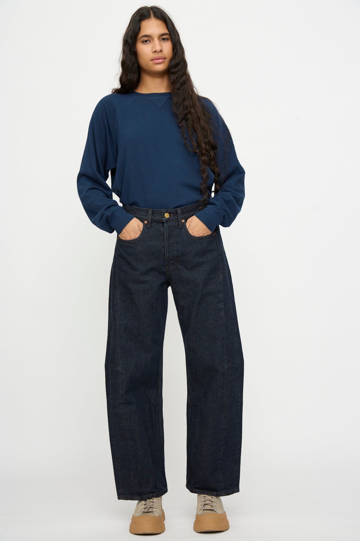 Person standing against a plain background, wearing a navy long-sleeve shirt and B Sides Slim Lasso in Indigo Brown Rinse jeans, with tan shoes and hands in pockets, hair down.