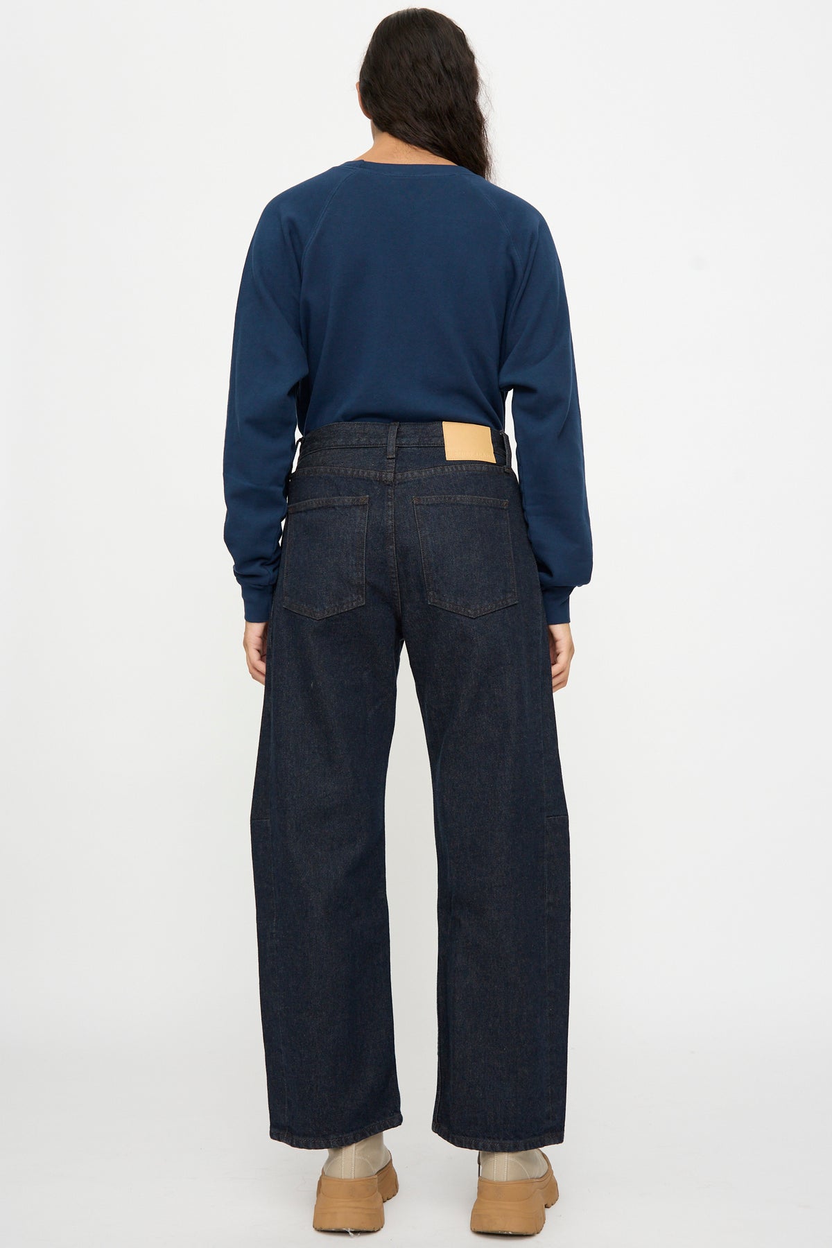 Person wearing a navy long-sleeve shirt, B Sides Slim Lasso in Indigo Brown Rinse jeans, and tan shoes, standing facing away against a white background.