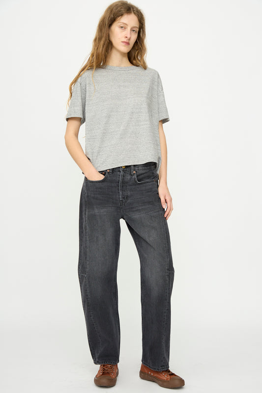 A person stands against a white background wearing a loose grey t-shirt, B Sides Slim Lasso Jean in Stil Black, and brown lace-up shoes, with one hand in a pocket.