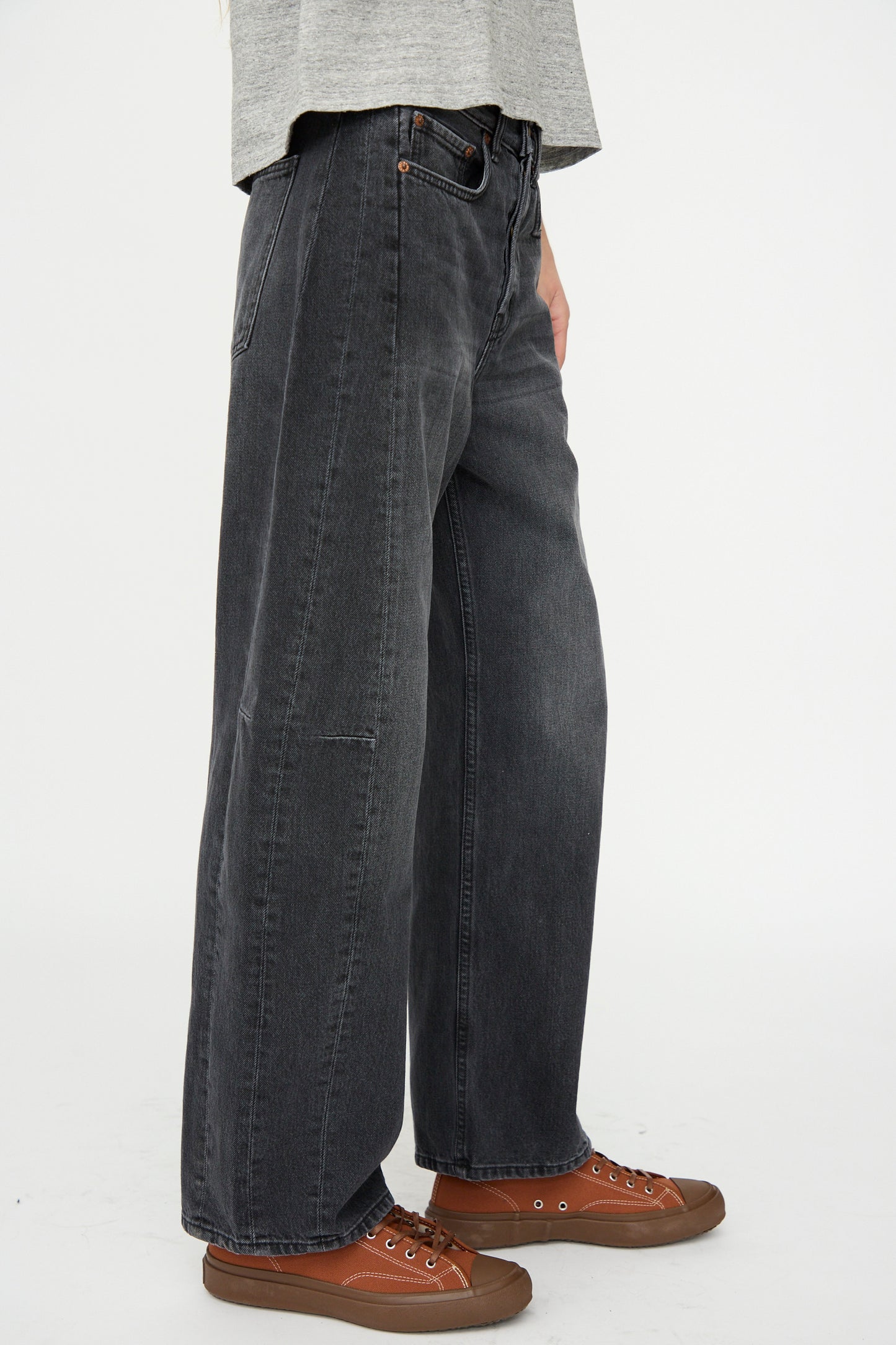 A person wears B Sides Slim Lasso Jean in Stil Black, paired with brown sneakers, standing against a plain white background. Only the lower torso and legs are visible.