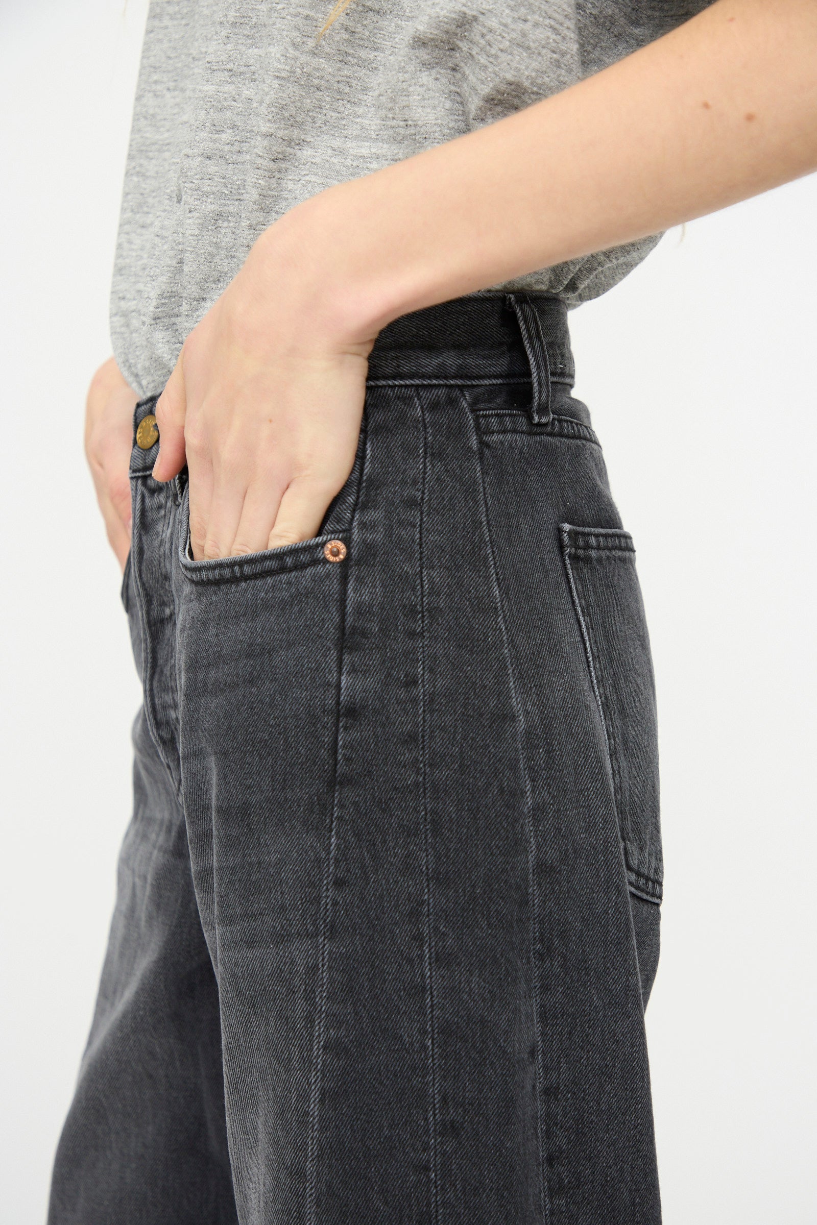 Person in a grey t-shirt and B Sides Slim Lasso Jean in Stil Black, with one hand in the front pocket, shown from mid-torso to upper thigh.