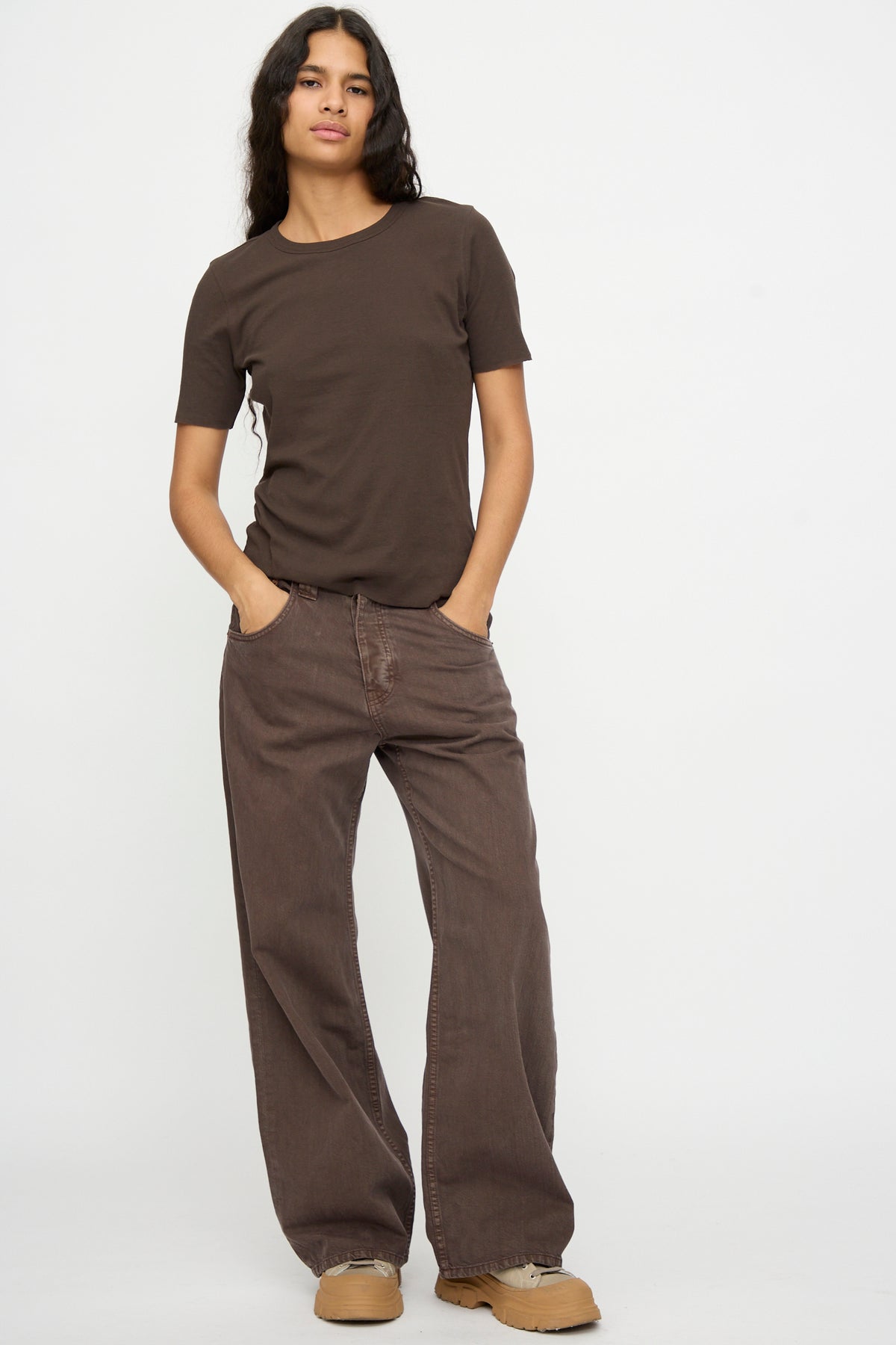 Person stands against a plain background wearing B Sides Vic Jean in Saddle Overdye, a brown t-shirt, tan shoes, and has hands in pockets.