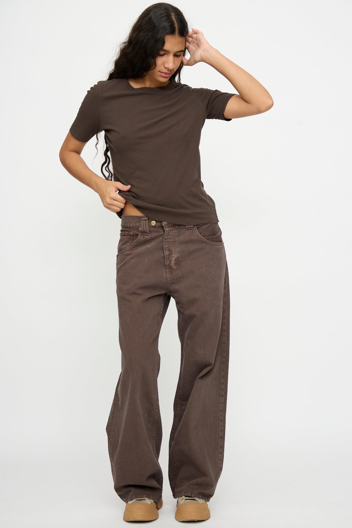 Person with long hair in a brown t-shirt, tan shoes, and B Sides Vic Jean in Saddle Overdye stands against a plain white background.