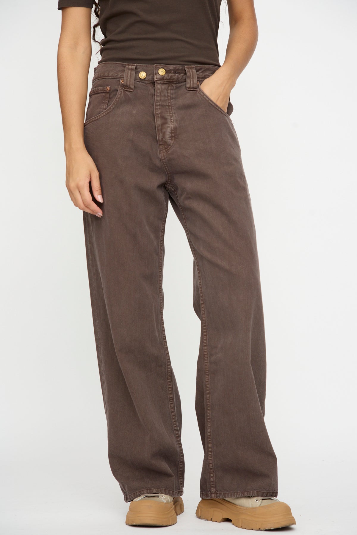 Person wearing B Sides Vic Jean in Saddle Overdye, a dark brown short-sleeve top, and tan shoes, standing with hands in pockets against a white background.