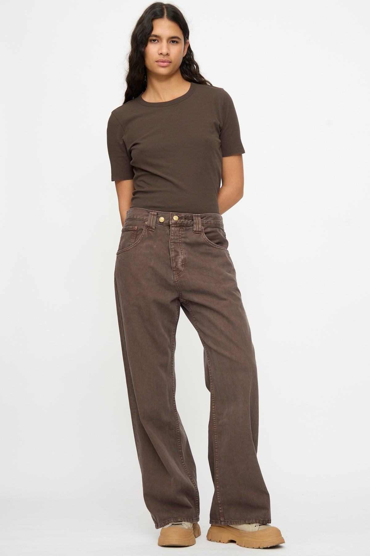 A person stands against a plain background wearing B Sides Vic Jean in Saddle Overdye, a brown short-sleeve t-shirt, tan shoes, with arms behind their back.