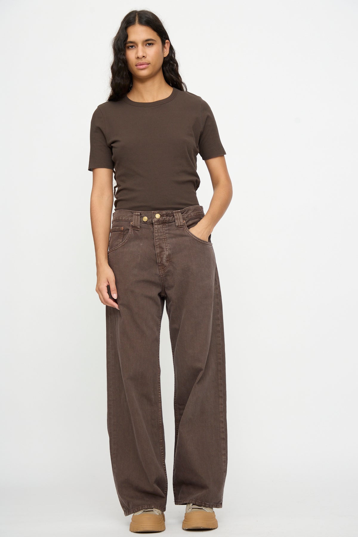 Person stands against a white background, wearing a brown short-sleeve shirt, tan shoes, and B Sides Vic Jean in Saddle Overdye with hands in pockets and a neutral expression.