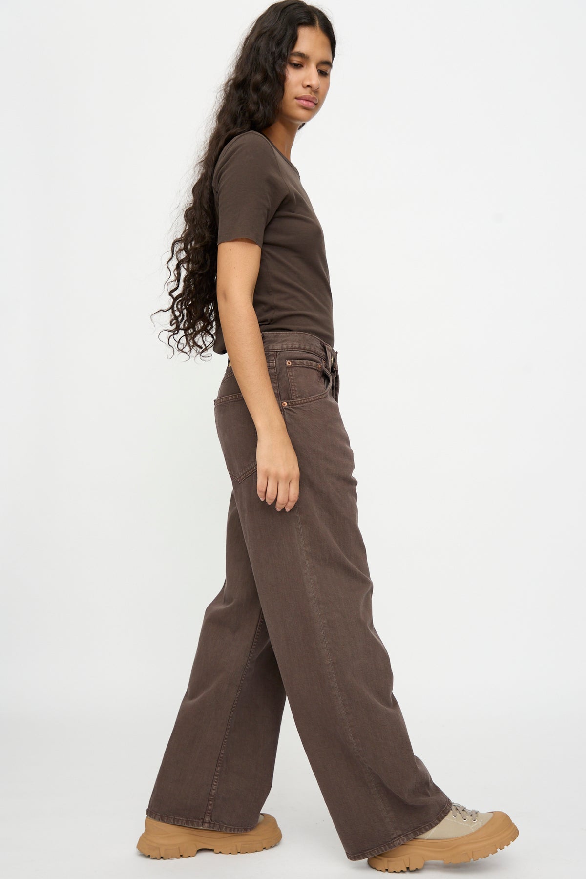 Person with long curly hair wearing a brown tee, Vic Jean in Saddle Overdye by B Sides, and tan sneakers, standing sideways against a white background.