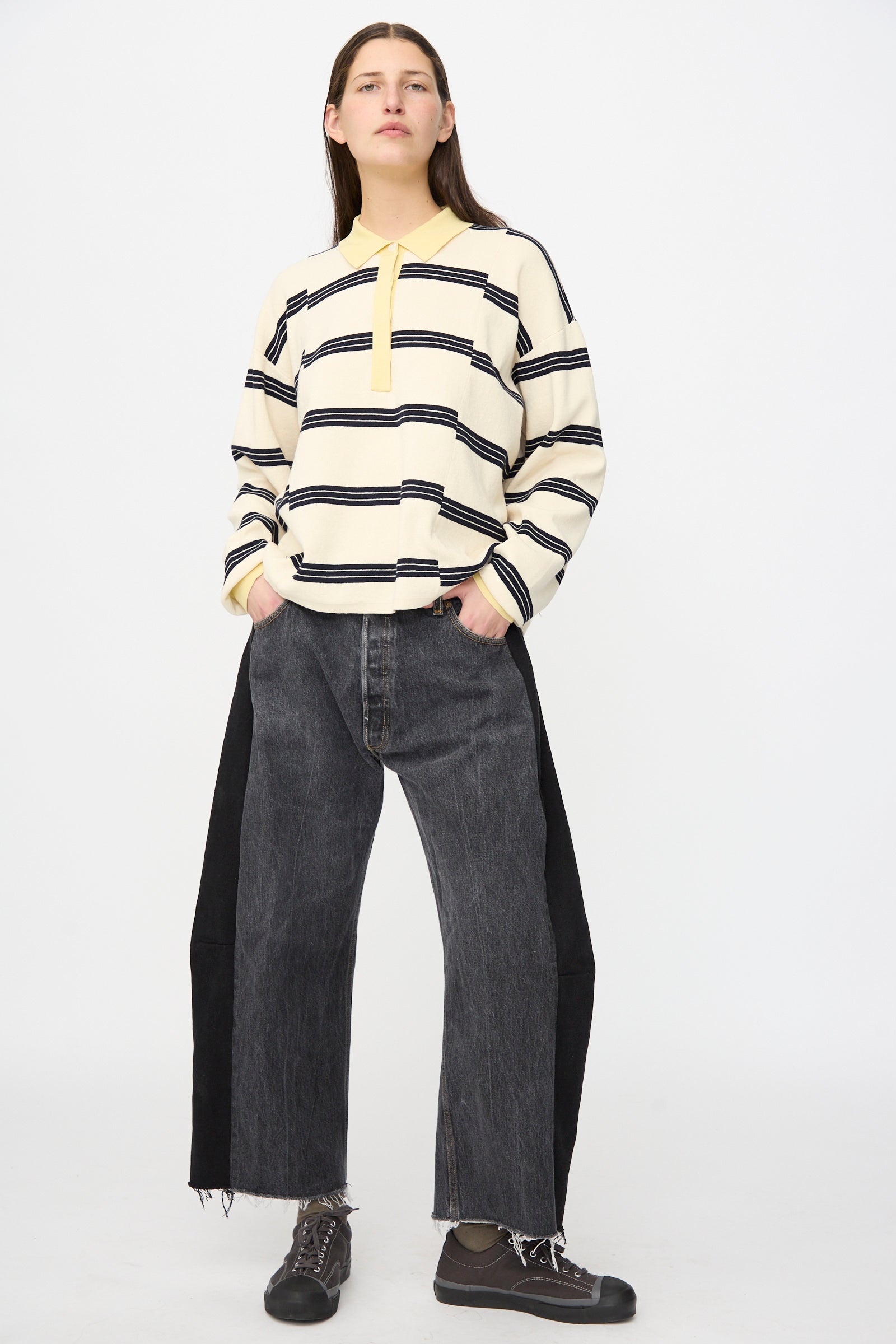 A person stands against a plain background wearing B Sides Vintage Lasso Jean in Black with raw hems, paired with a striped long-sleeve shirt and dark shoes. Their hands are in their pockets, and they have a neutral expression.