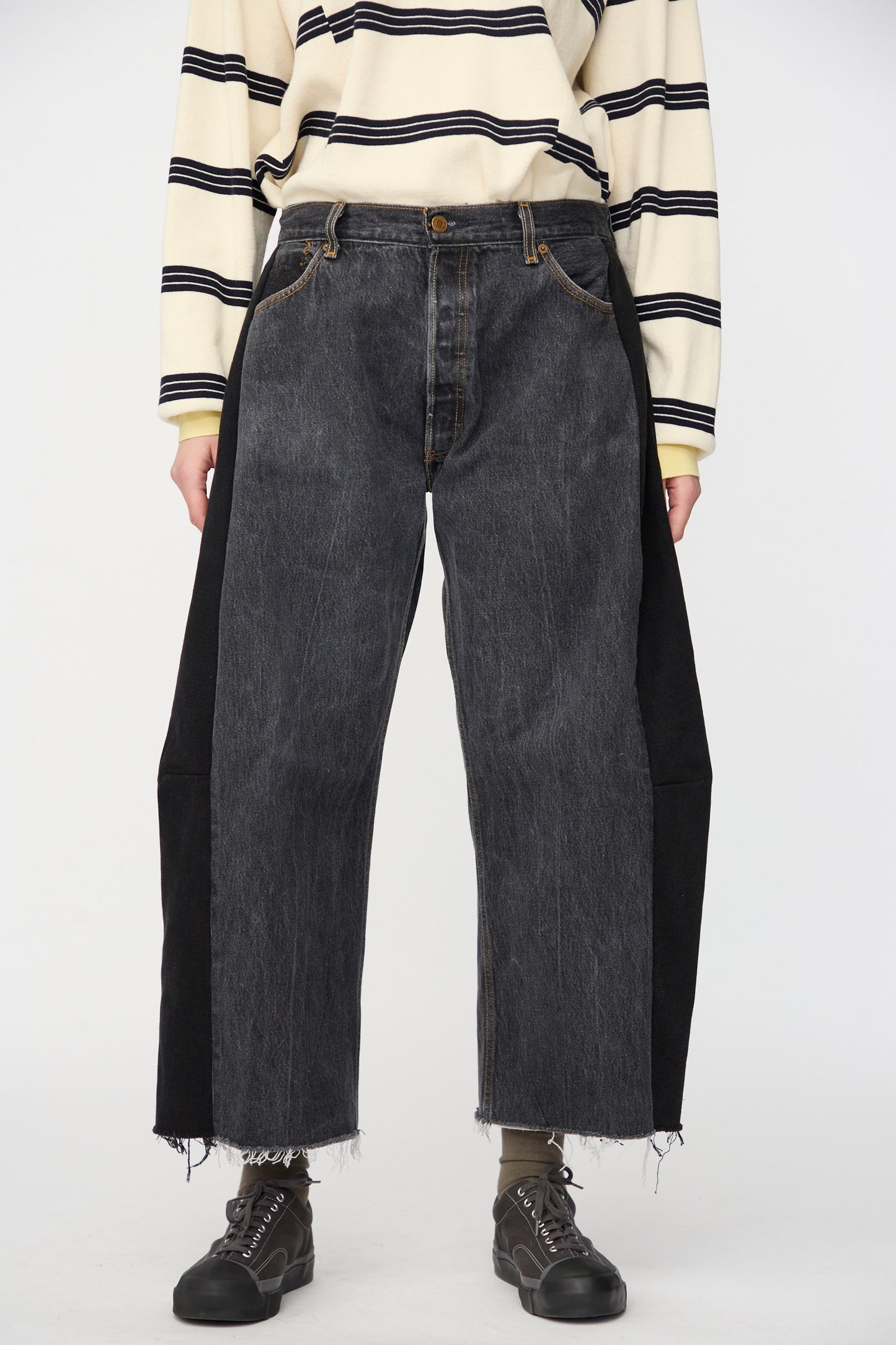 A person wears a striped long-sleeve shirt, B Sides Vintage Lasso Jean in Black with frayed hems, and black shoes, standing against a plain background.