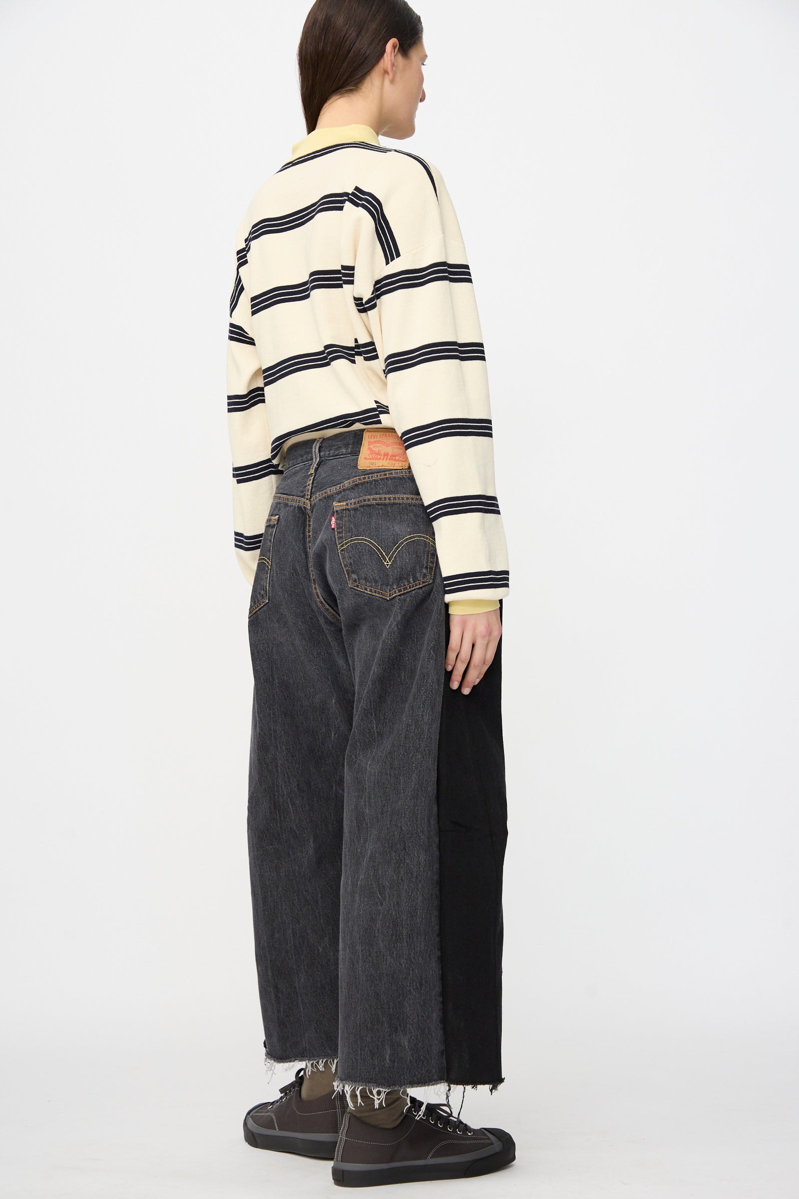 A person stands in profile against a white background, wearing the B Sides Vintage Lasso Jean in Black and a beige and black striped long-sleeve shirt with dark sneakers.