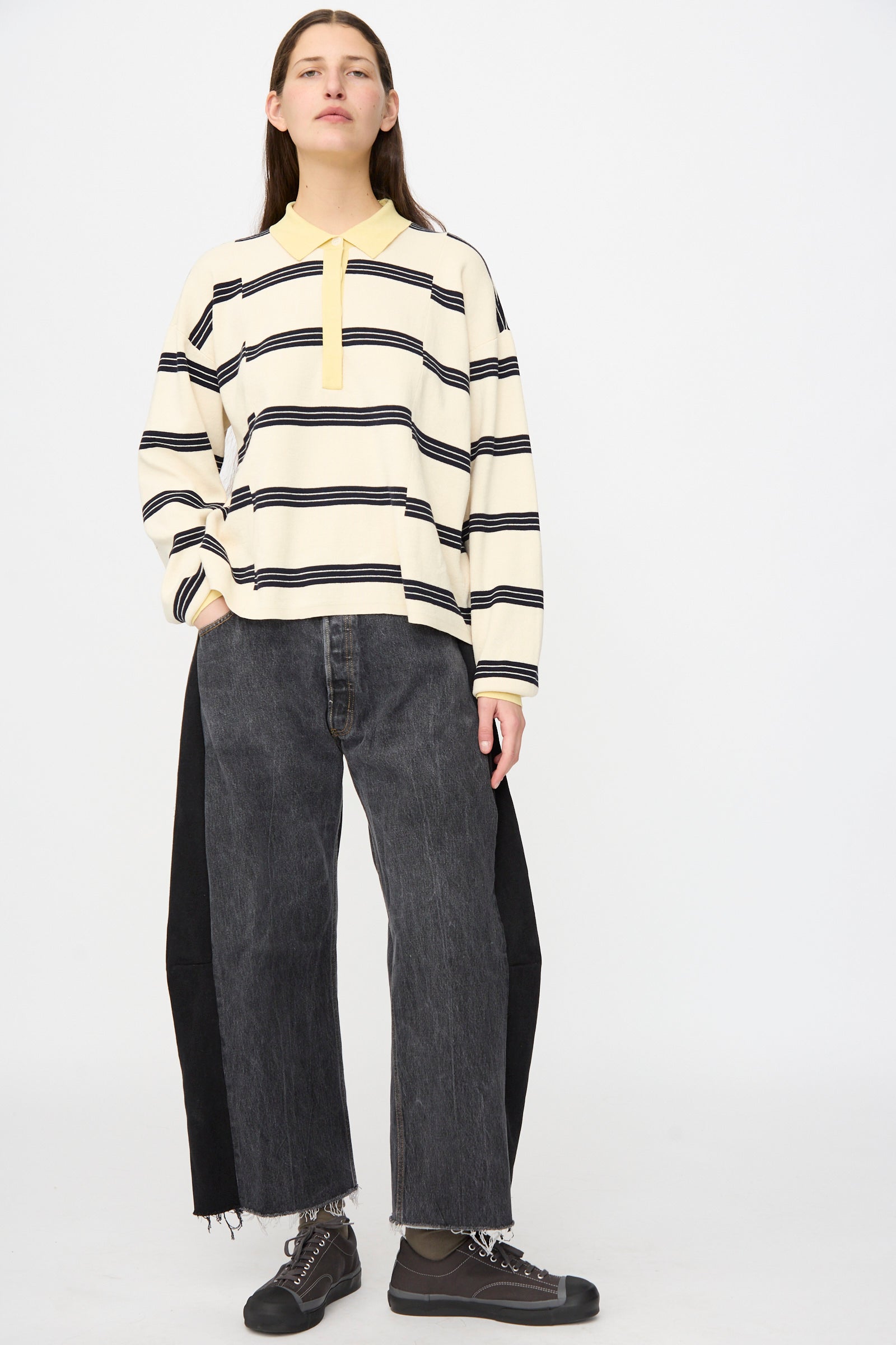 Person stands against a plain background, wearing a cream and black striped long-sleeve shirt, B Sides Vintage Lasso Jean in Black with relaxed fit and frayed hems, paired with dark sneakers.