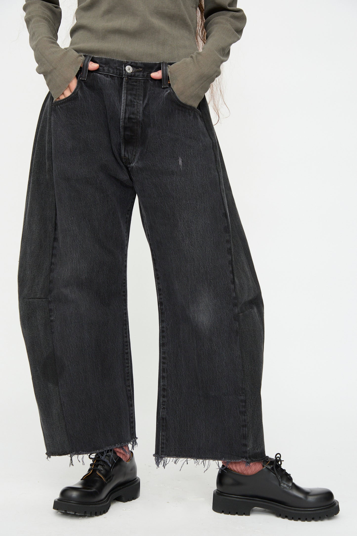A person wears B Sides Vintage Lasso Jean in Black, a long-sleeve olive shirt, and black chunky shoes, standing against a plain white background.