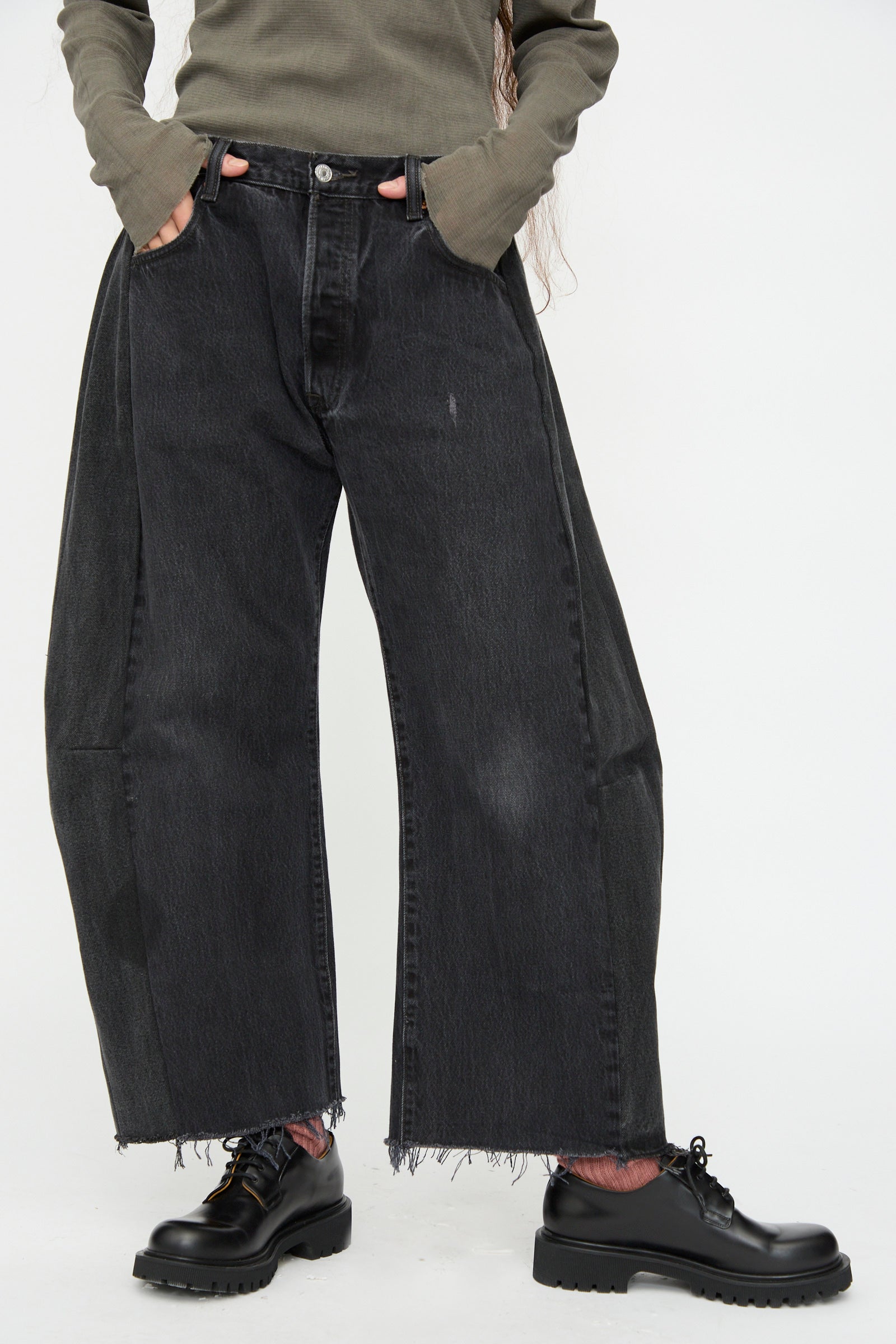 A person wears B Sides Vintage Lasso Jean in Black, a long-sleeve olive shirt, and black chunky shoes, standing against a plain white background.