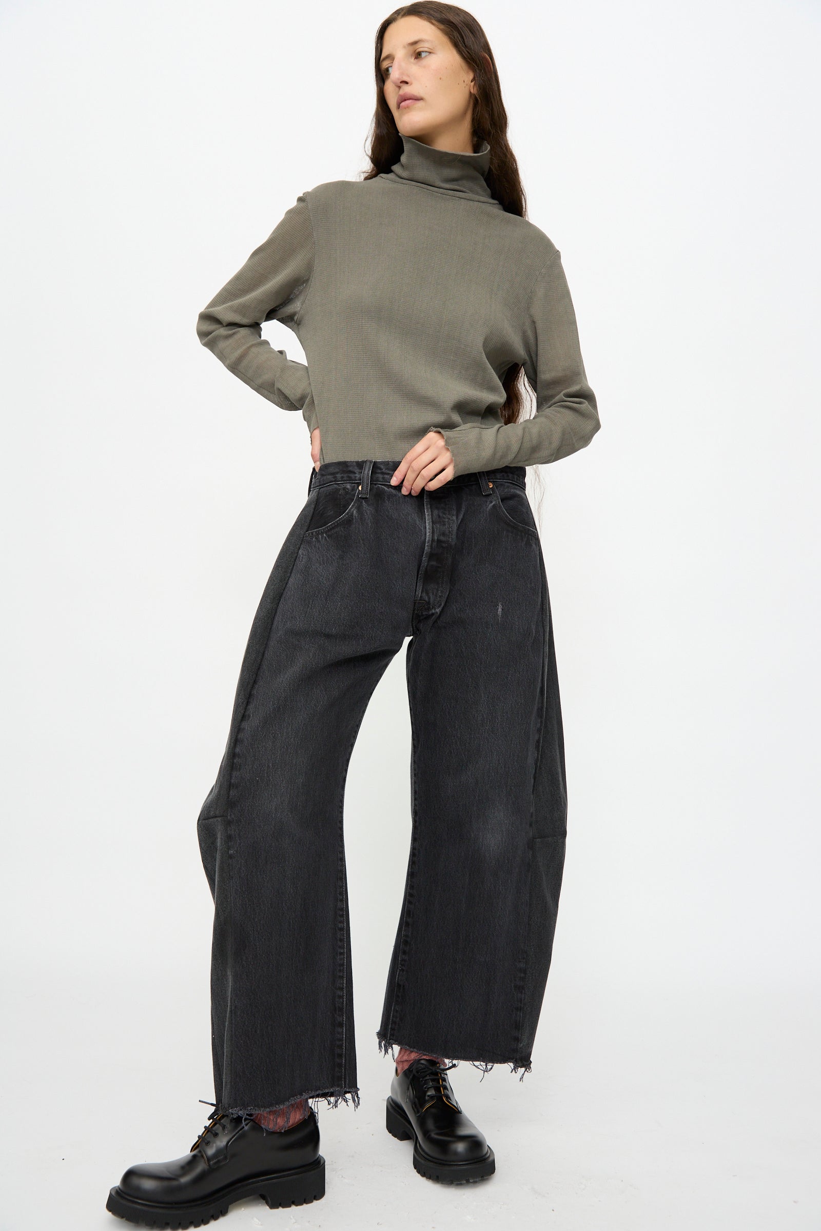 A person stands with one hand on hip, wearing a grey turtleneck sweater, B Sides Vintage Lasso Jean in Black, and black lace-up shoes against a plain white background.