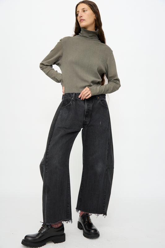 A person stands with one hand on hip, wearing a grey turtleneck sweater, B Sides Vintage Lasso Jean in Black, and black lace-up shoes against a plain white background.
