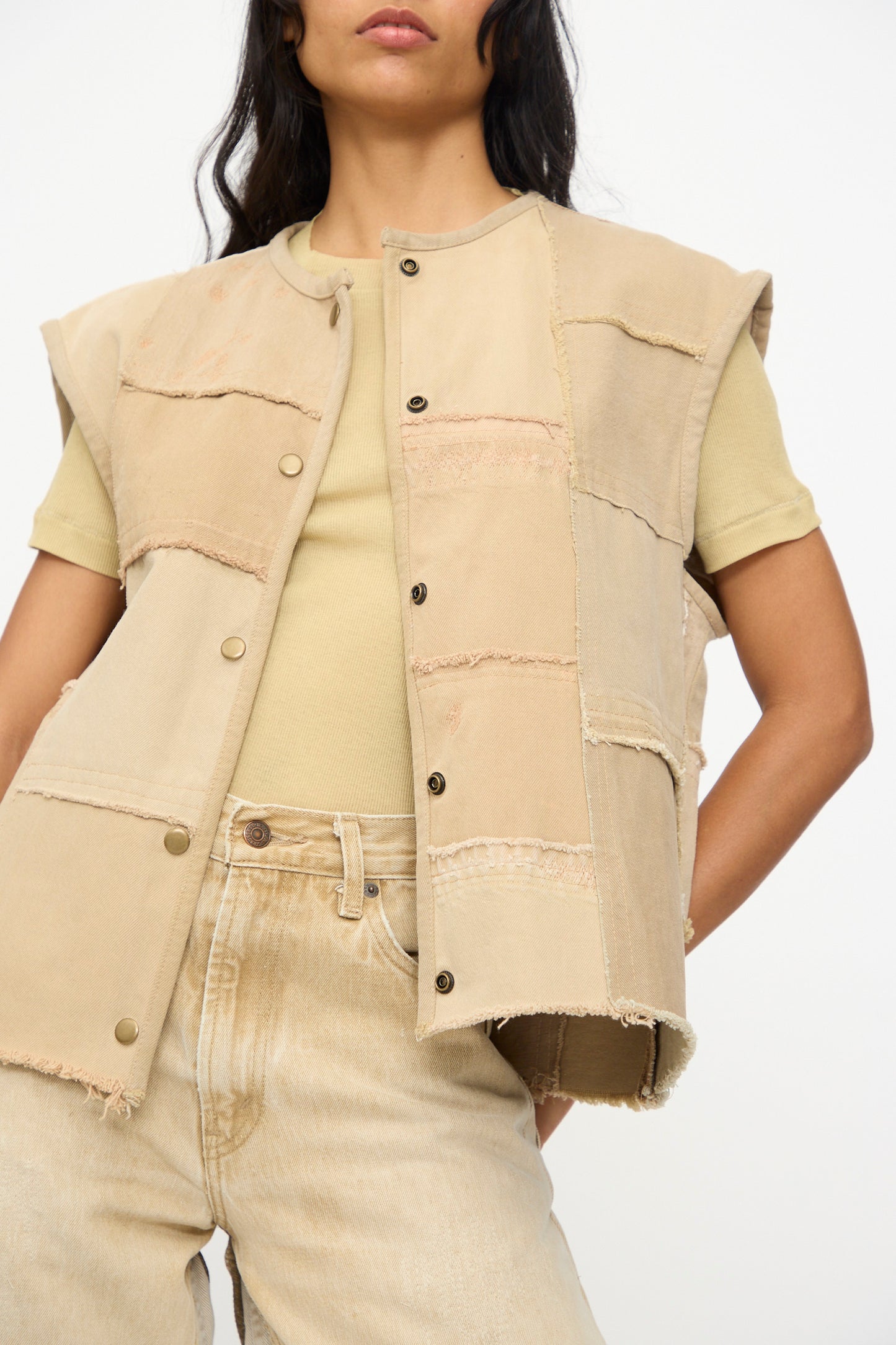 A person wears the B Sides Vintage Madras Vest in Khaki over a beige t-shirt and light jeans, standing against a plain background. The vest brings vintage charm to the minimal outfit.