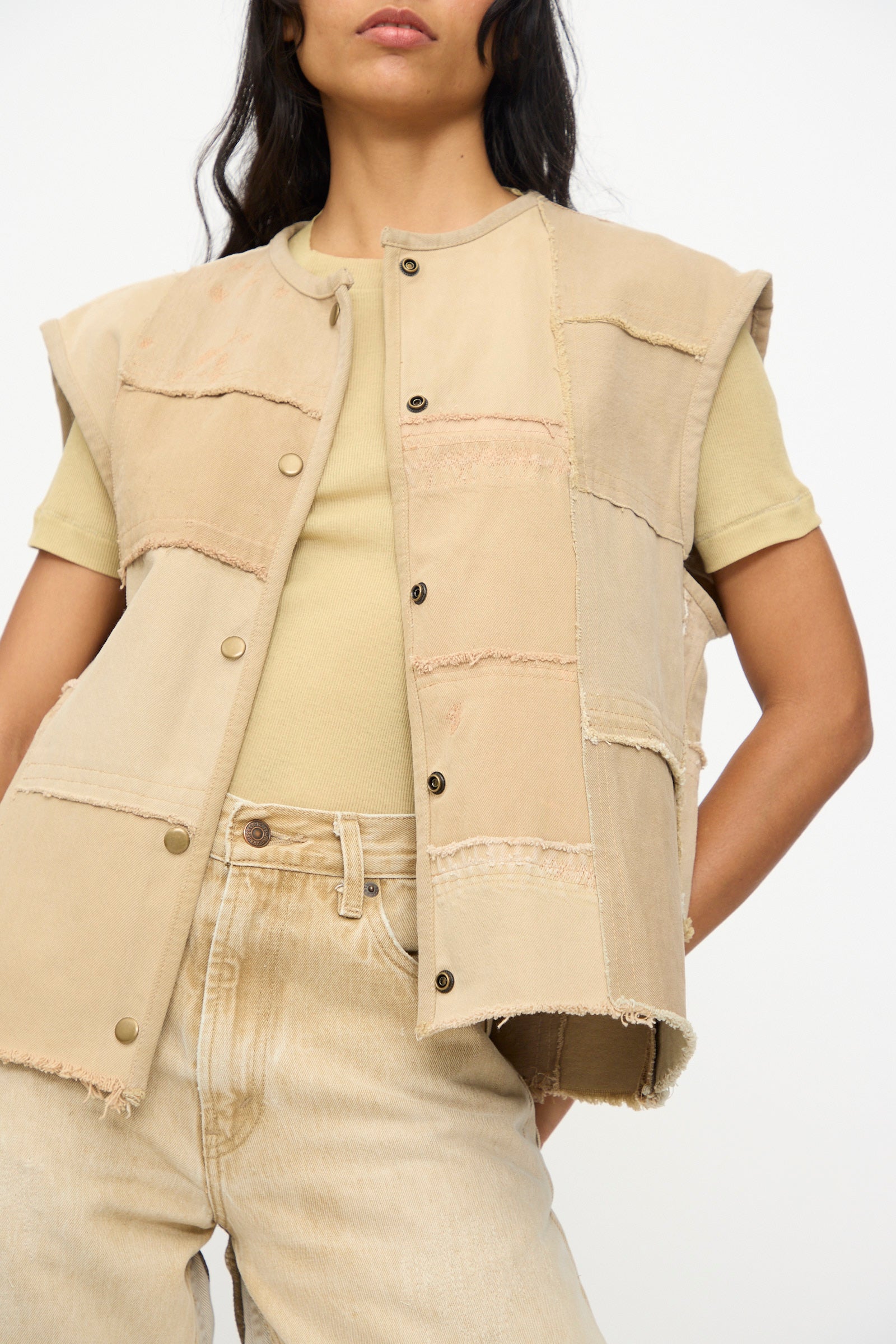 A person wears the B Sides Vintage Madras Vest in Khaki over a beige t-shirt and light jeans, standing against a plain background. The vest brings vintage charm to the minimal outfit.