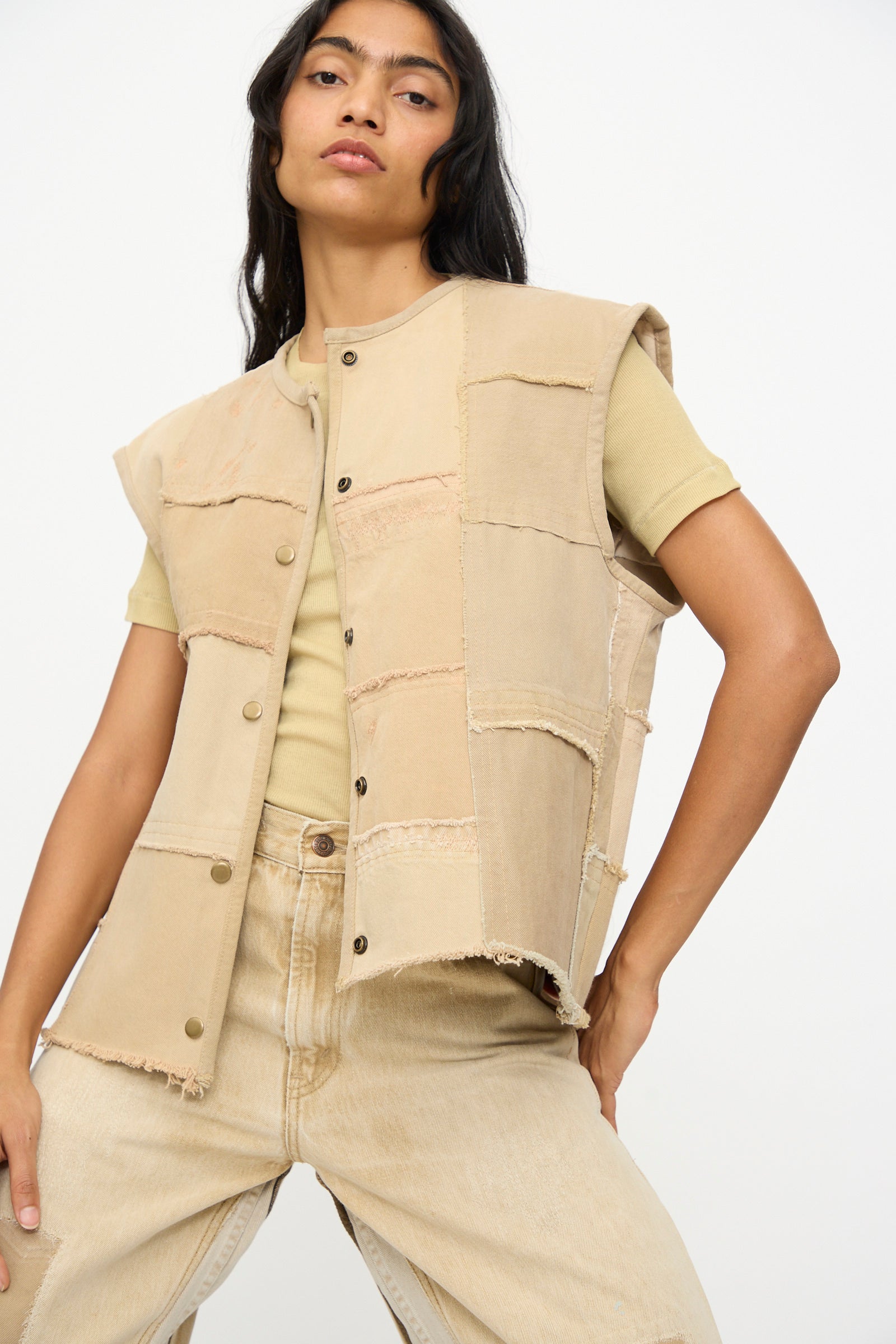 Model wears the B Sides Vintage Madras Vest in Khaki over a light yellow t-shirt and beige jeans, posing with hand on hip against a plain background.