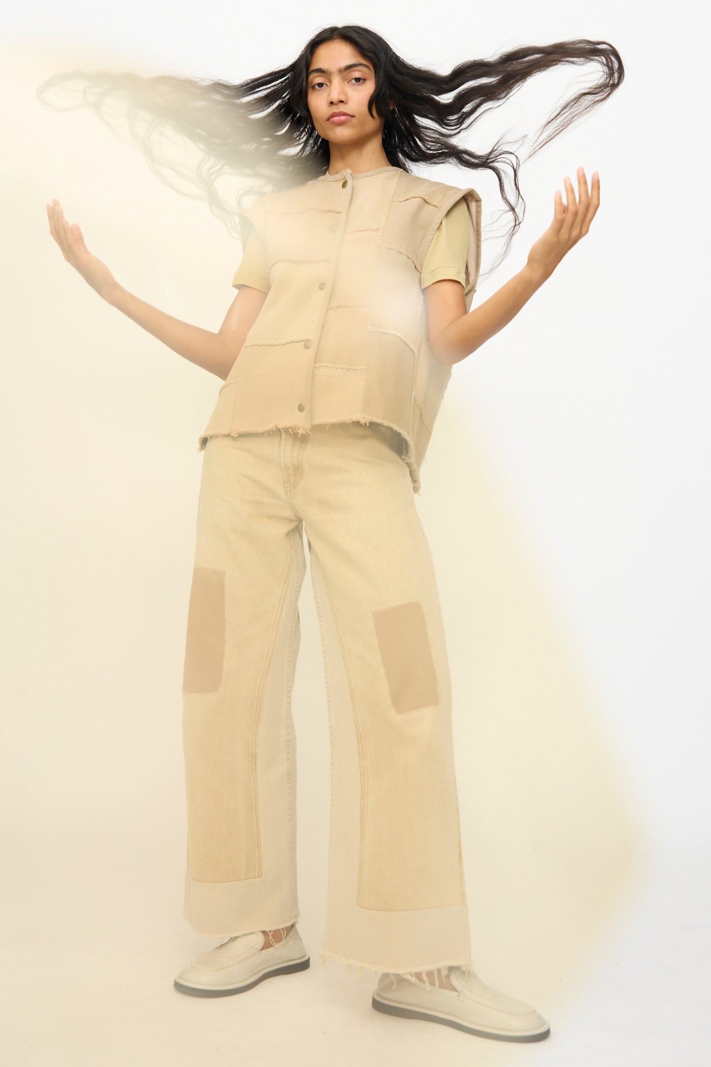 A person stands with arms raised, wearing the B Sides Vintage Madras Vest in Khaki and wide-leg pants with patch details, paired with light-colored shoes, against a plain light background.