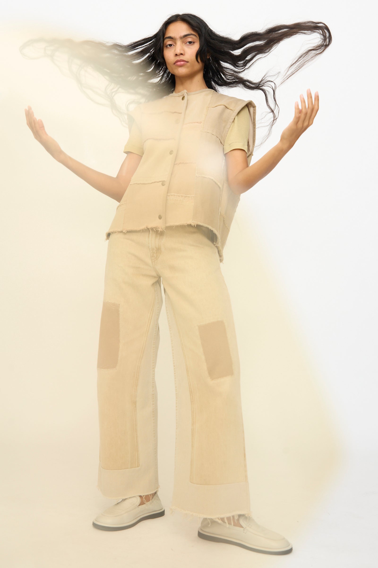 A person stands with arms raised, wearing the B Sides Vintage Madras Vest in Khaki and wide-leg pants with patch details, paired with light-colored shoes, against a plain light background.