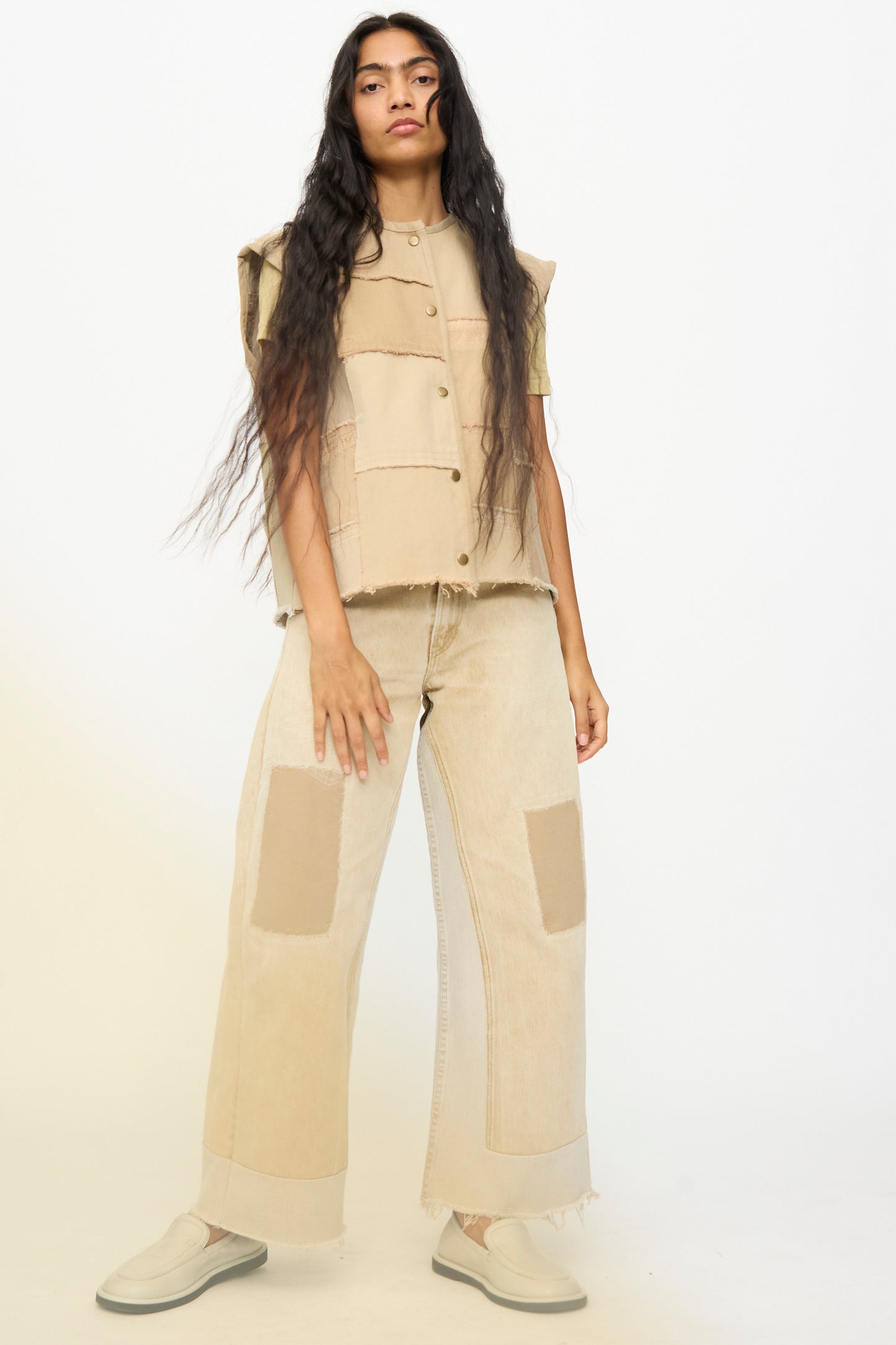 A person with long dark hair wears the Vintage Madras Vest in Khaki by B Sides and wide-leg patchwork pants, paired with slip-on shoes, standing against a plain light background.